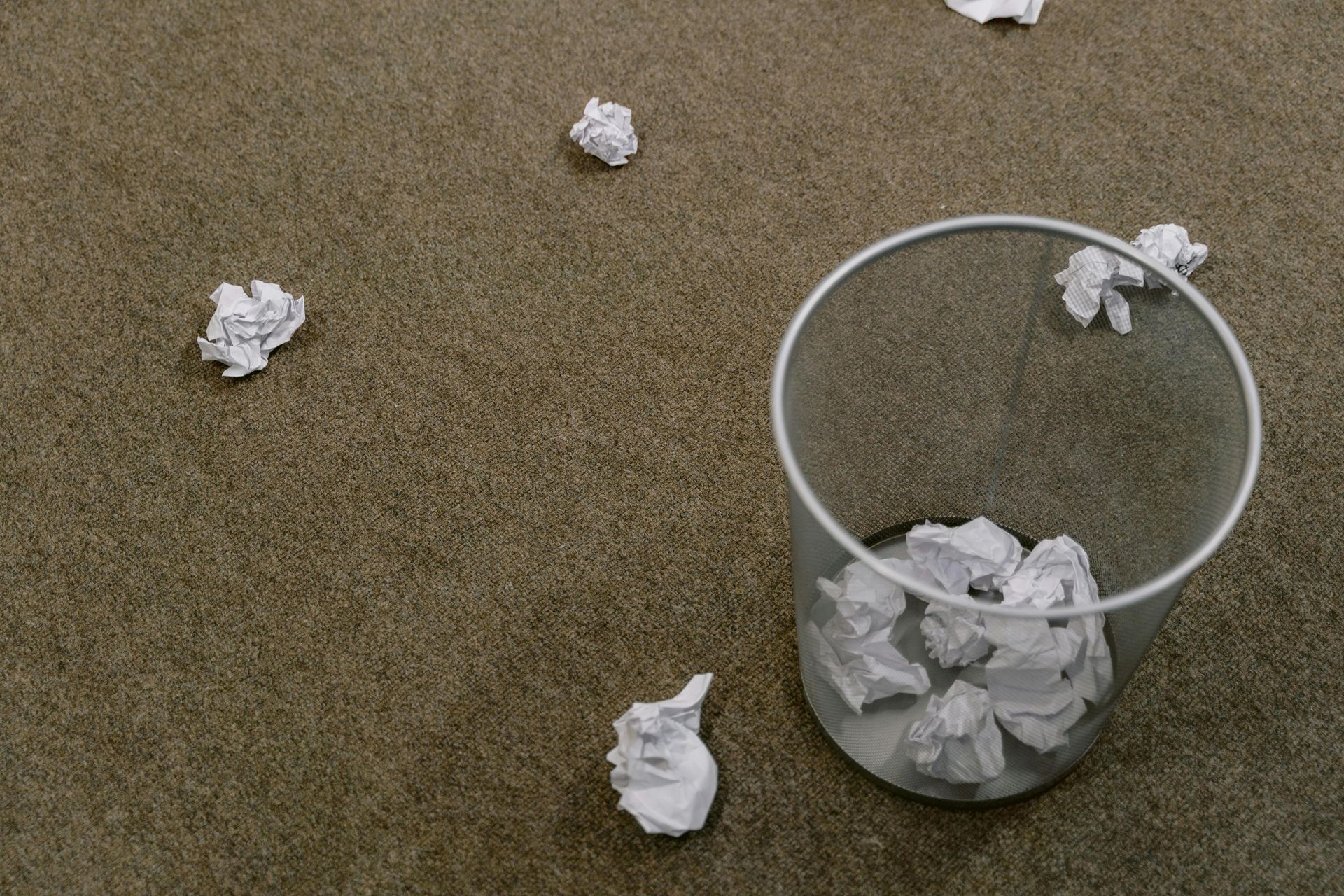Several crumpled pieces of white paper are scattered on a brown carpet around a small, wire-mesh wastebasket.