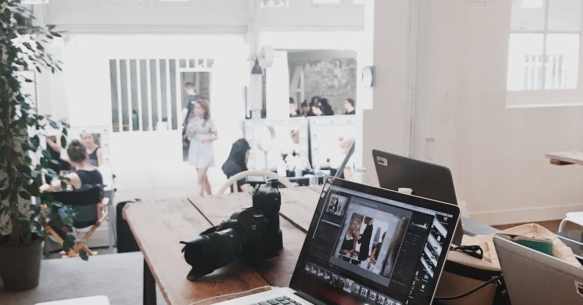 Laptop and camera on wooden desk, open office with people working and a woman walking by.