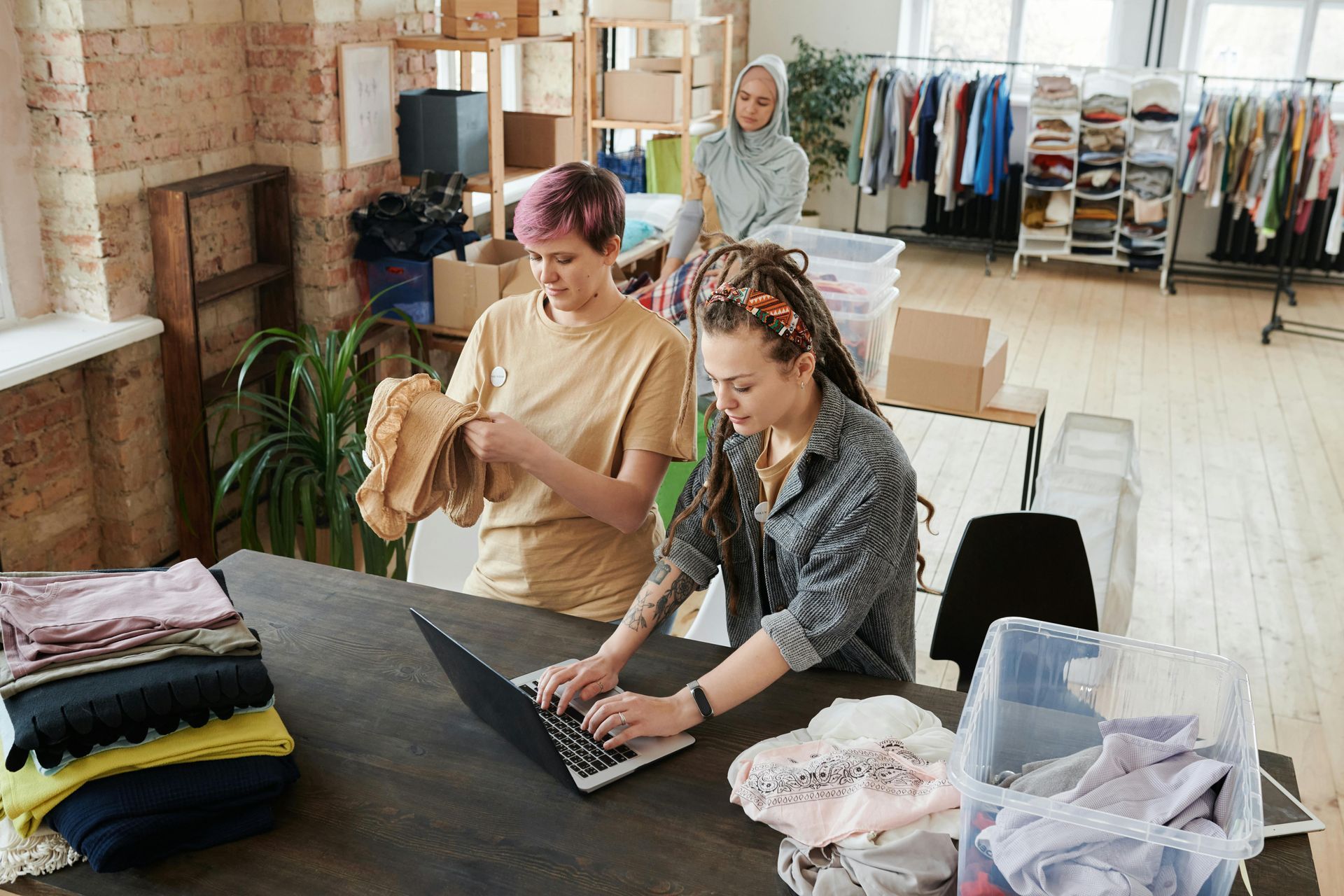 Three people sorting and organizing clothing in a bright room with a laptop on a table.