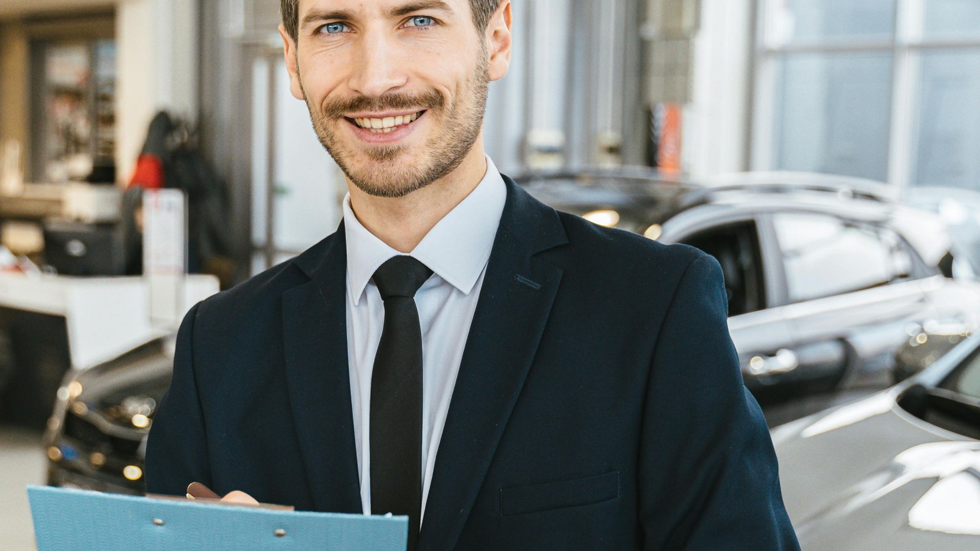 Man in suit smiles, holding clipboard, in a car dealership.