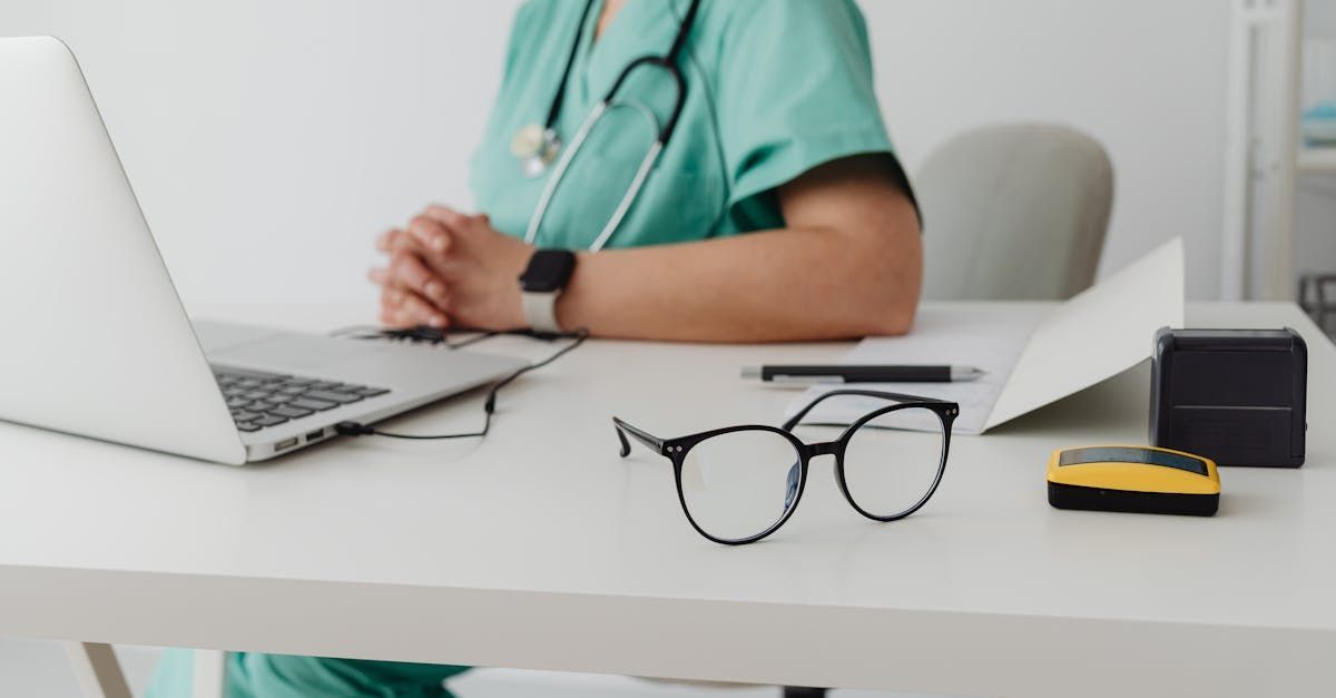 Doctor in scrubs at desk with laptop, glasses, and medical equipment.