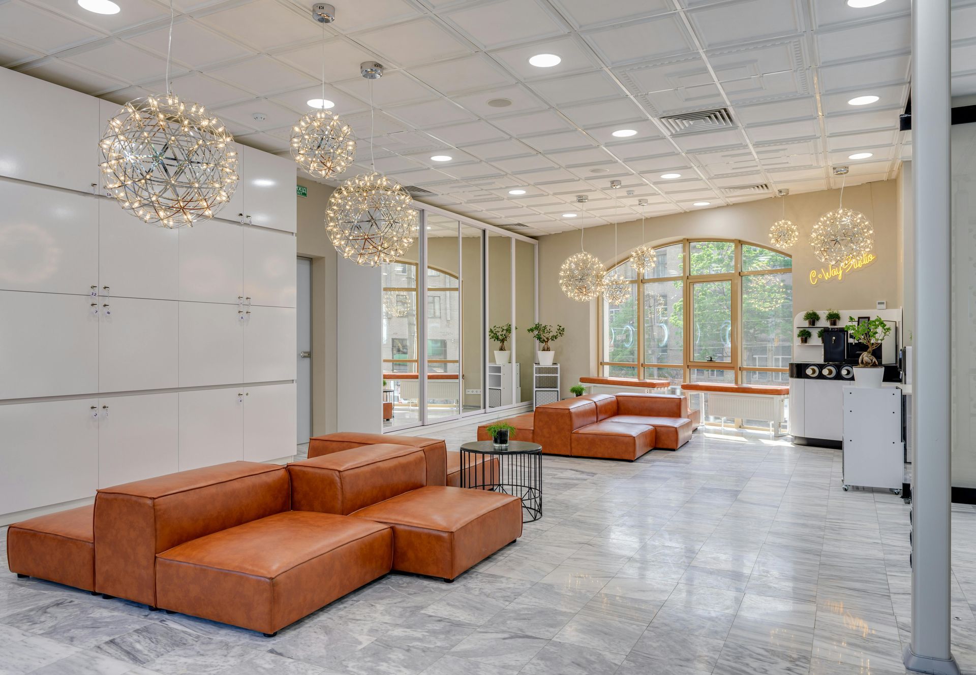 Lounge area with orange modular sofas, globe chandeliers, and large windows. White storage cabinets on the left.