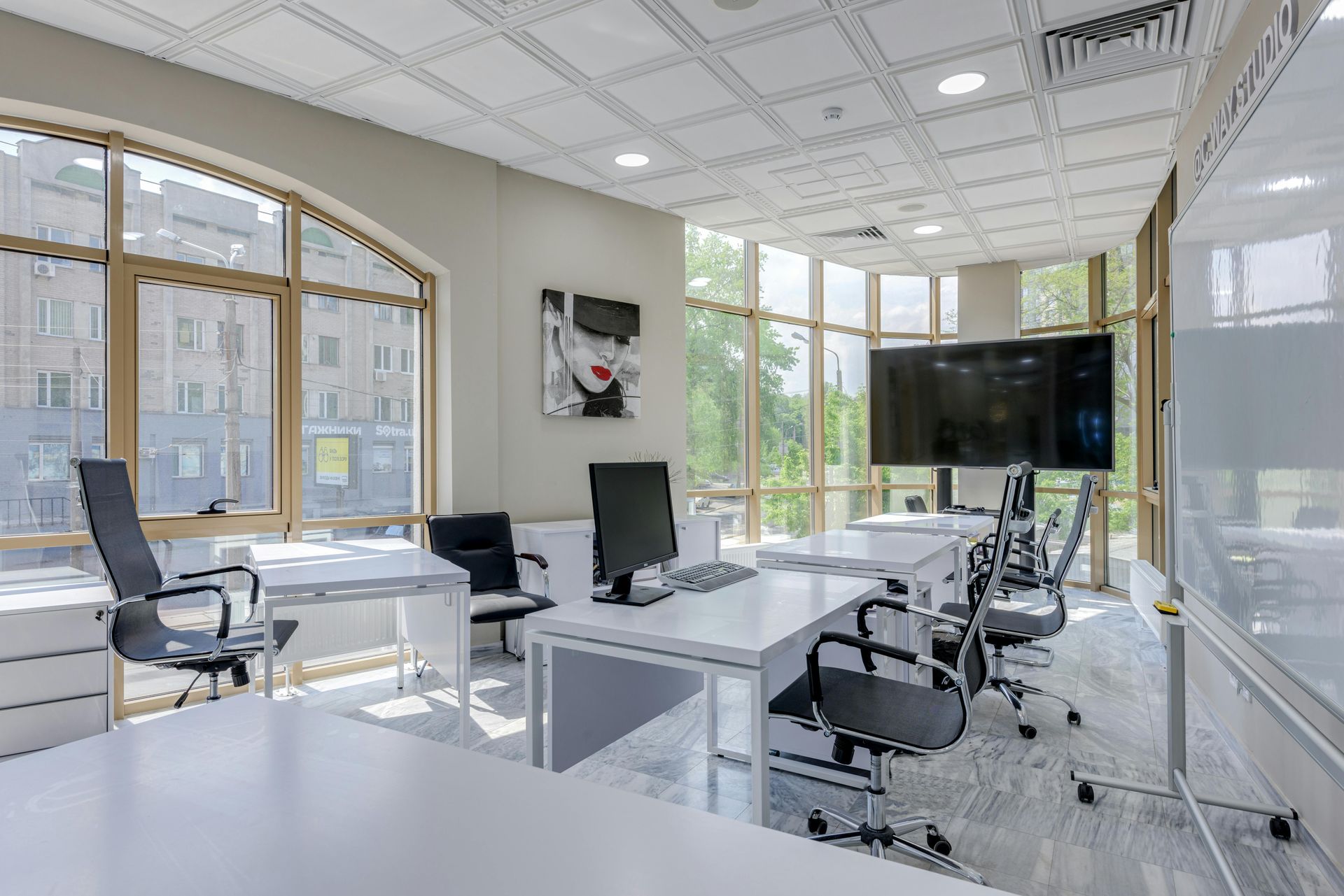Modern office with large windows, white desks, black chairs, a flat-screen TV, and art.