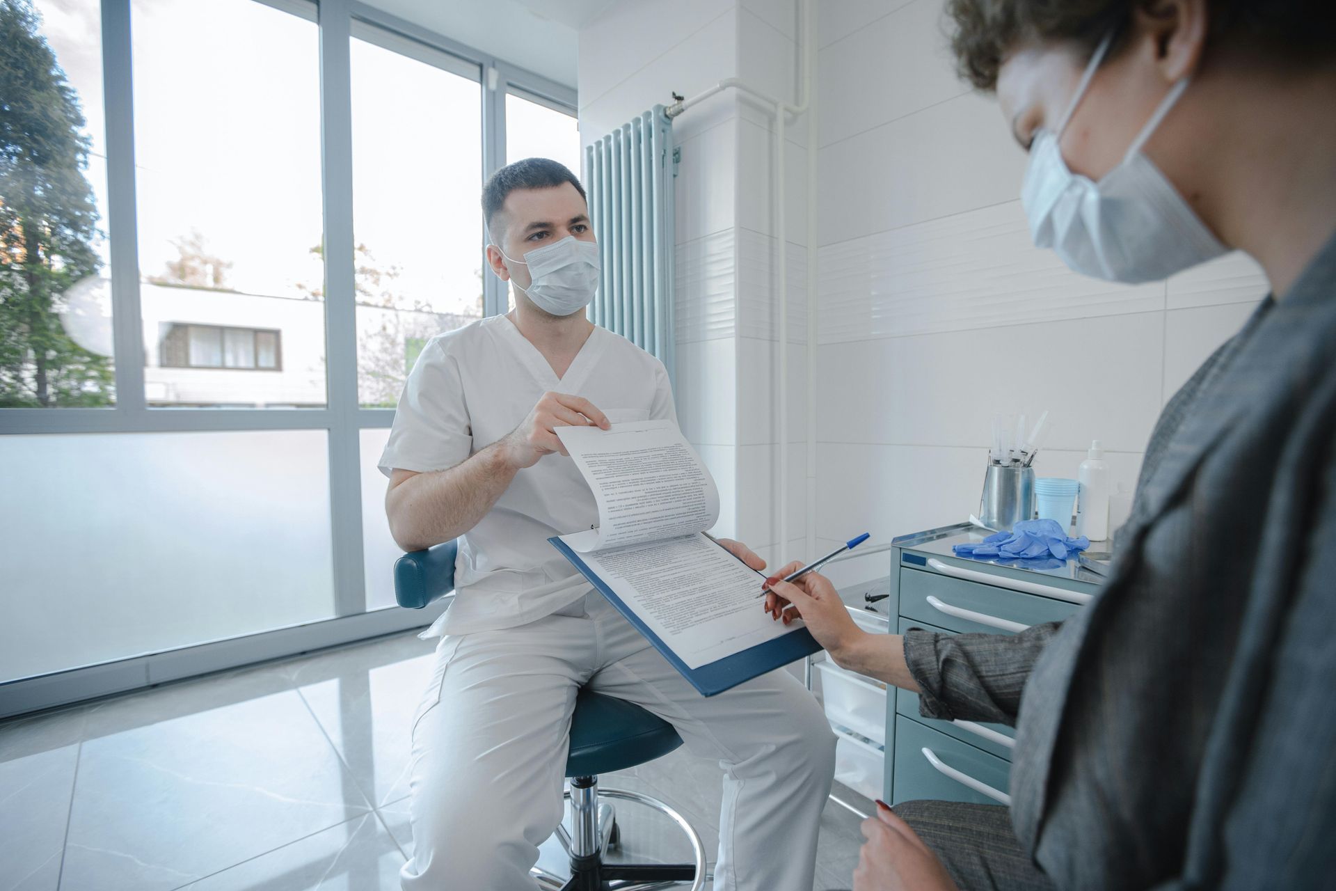 Doctor in mask shows paperwork to patient, who is also masked, in a bright medical office.