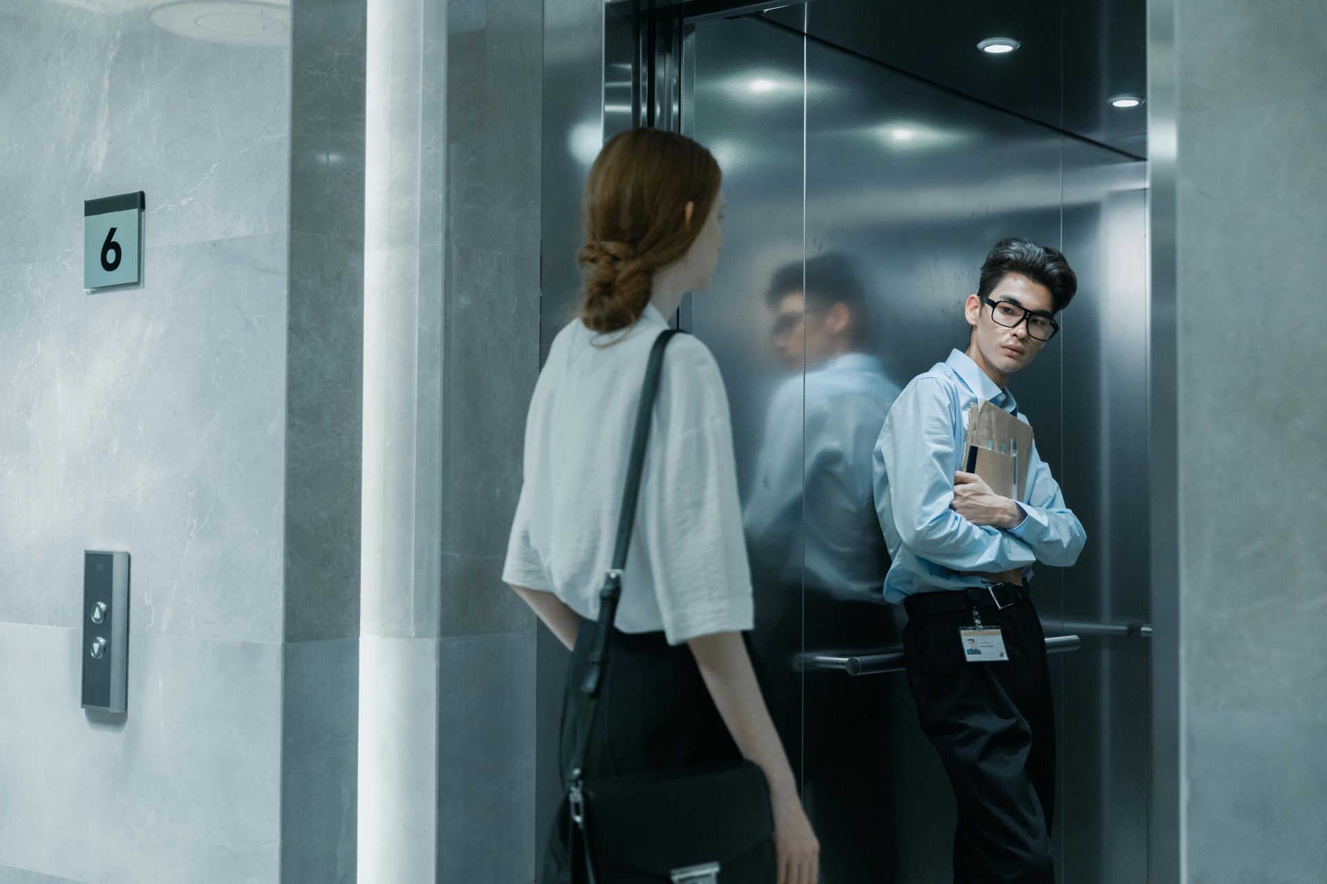 Woman and man in an elevator. Man in glasses, holding file, looking at the woman. Grey and white interior.