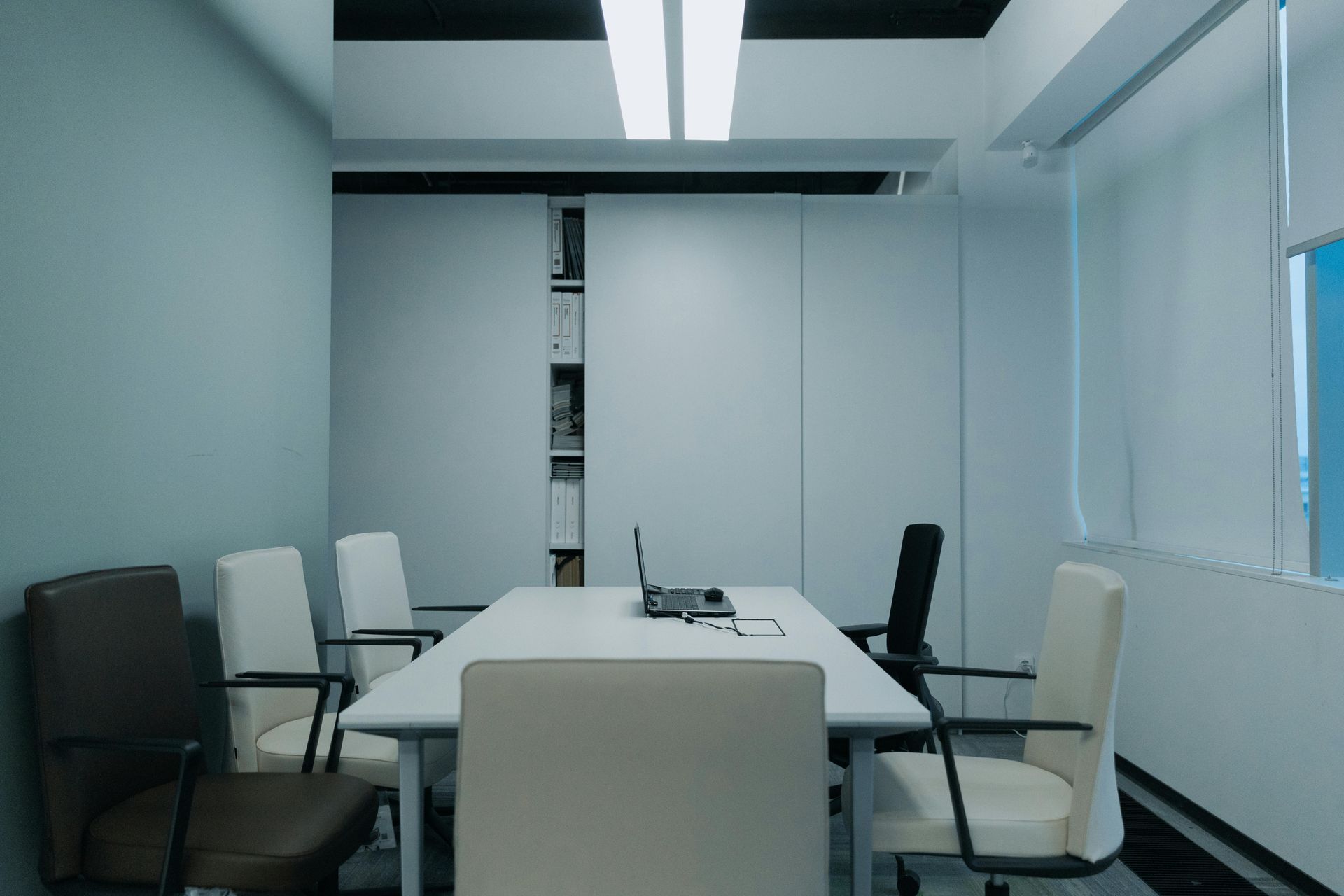 Meeting room with a white table, chairs, laptop, and built-in shelving. Pale blue walls and bright lighting.