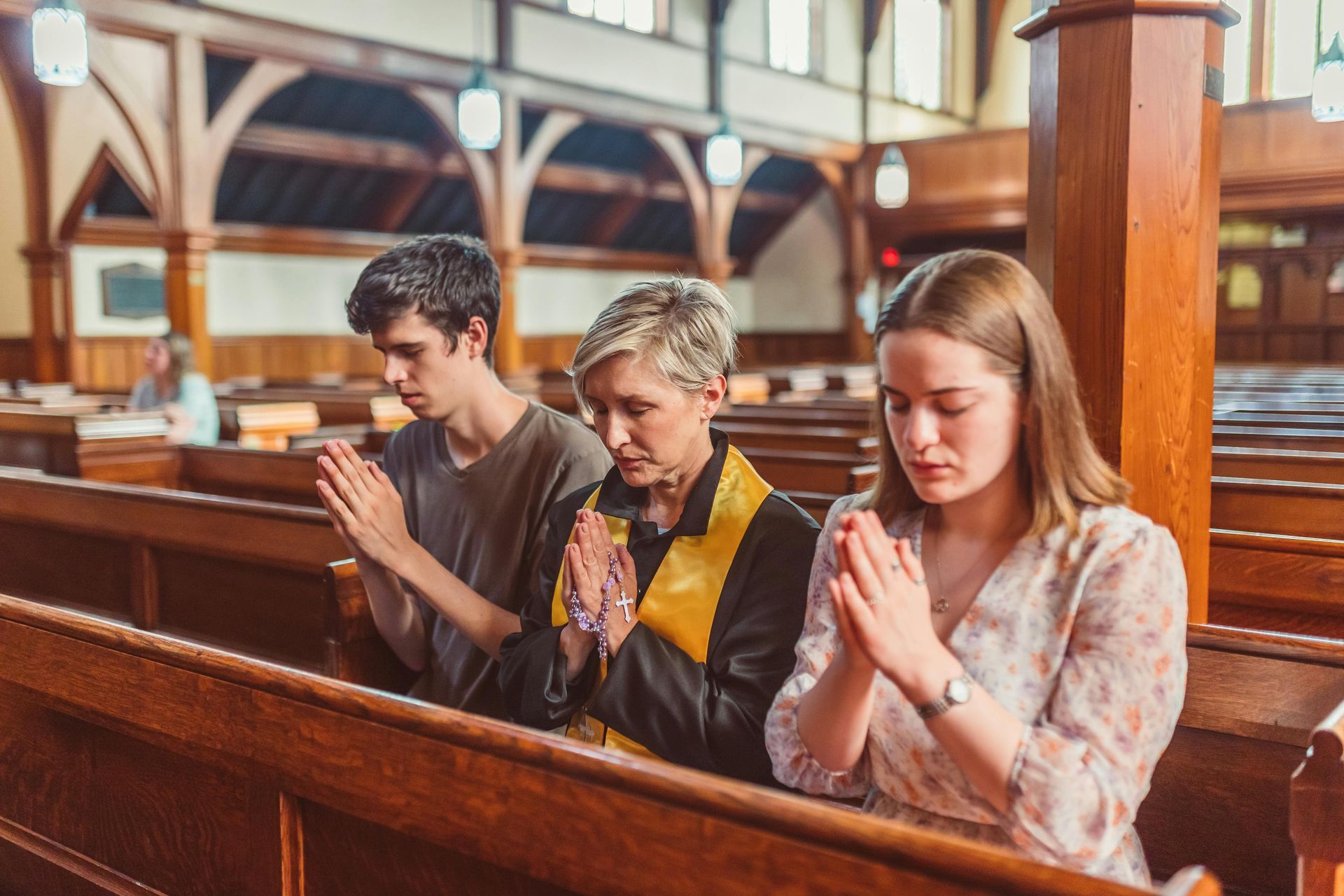 Three people praying in a church pew; hands clasped, heads bowed. Wooden interior, natural light.