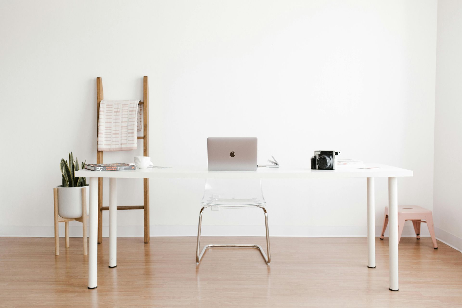 Minimalist white office with laptop on desk, clear chair, wooden ladder and plant.