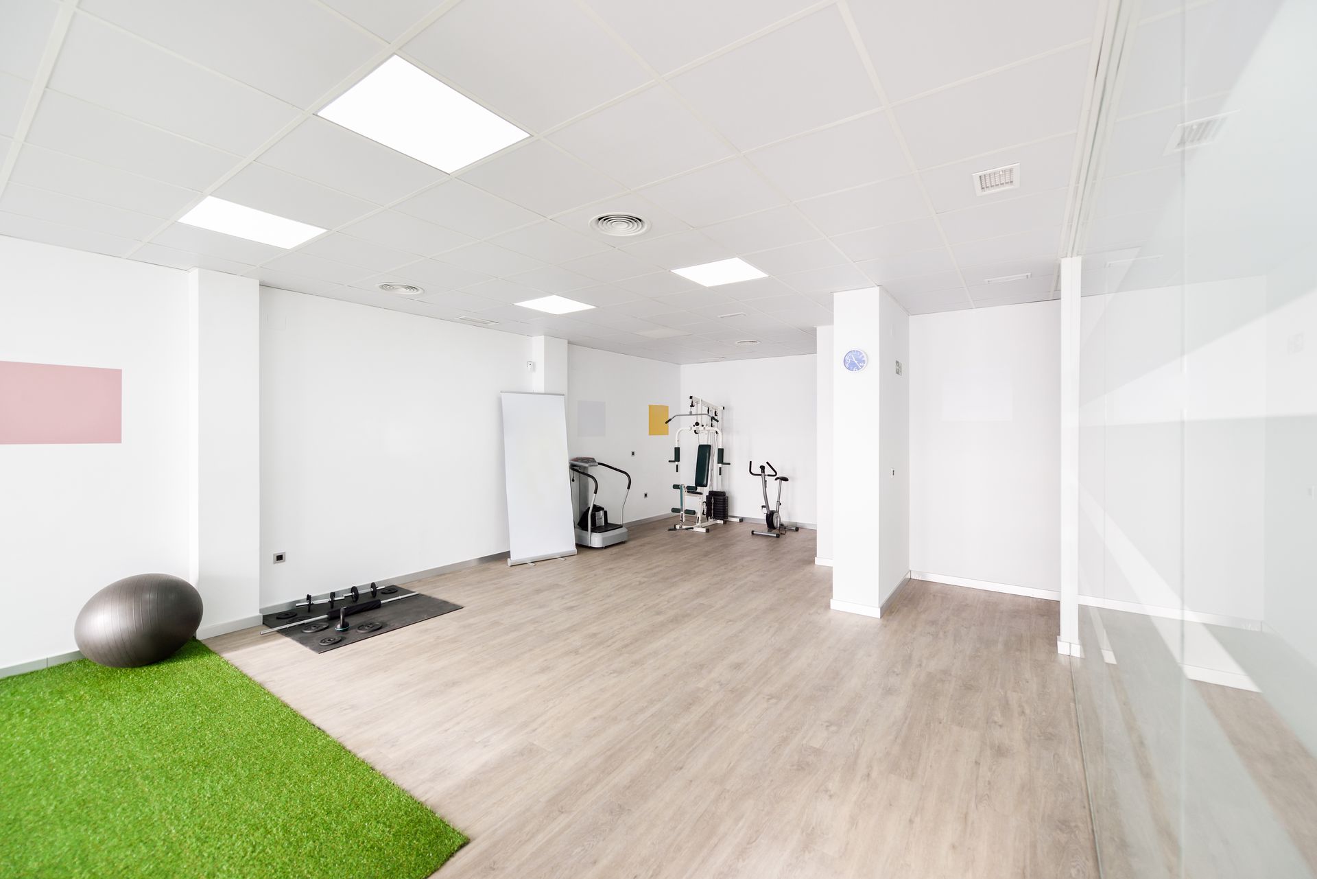 Empty gym with exercise equipment: weights, ball, machine, and green turf area on the floor.
