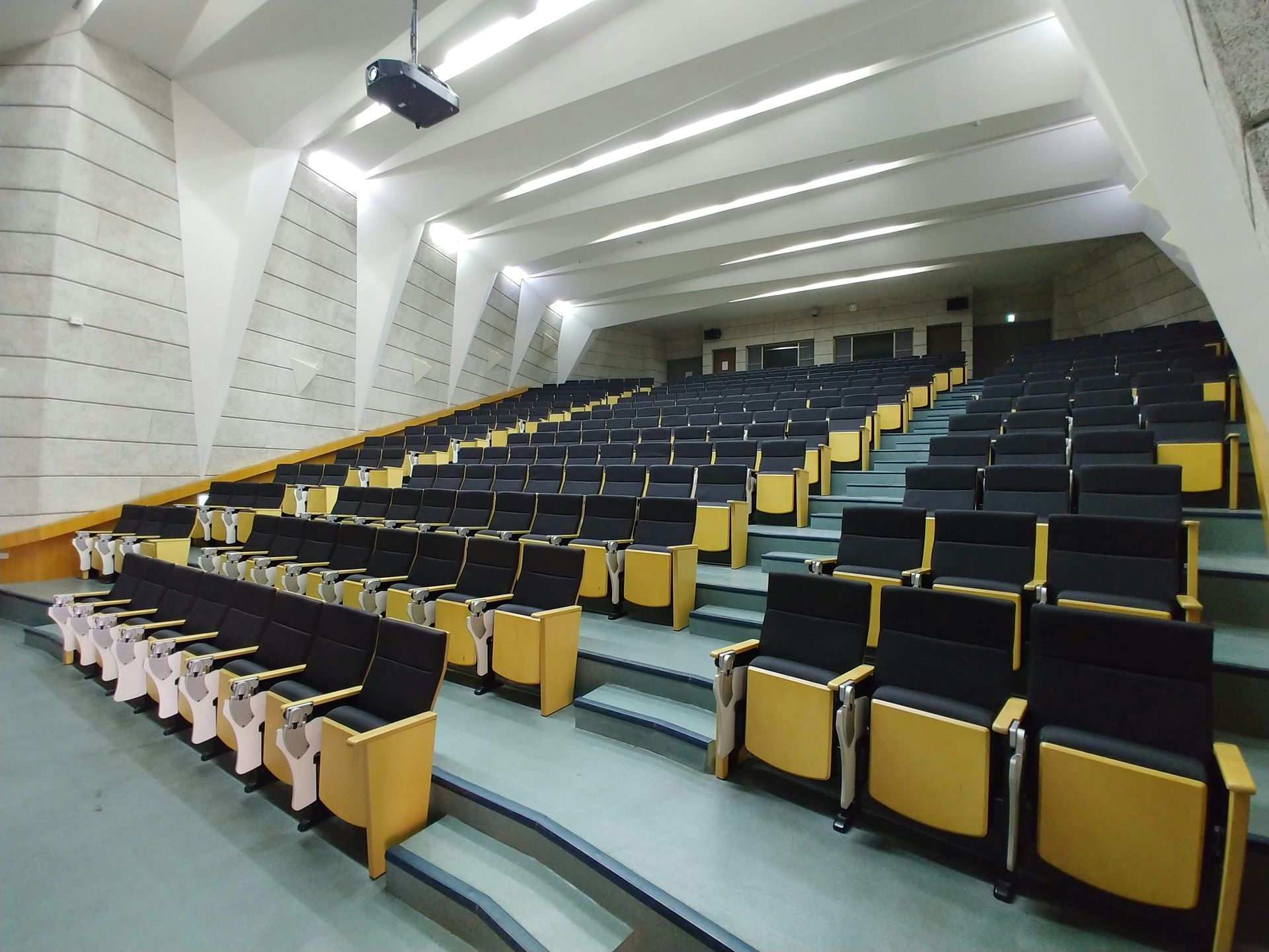 Auditorium with rows of black seats, yellow armrests, and angled walls, a projector hangs from the ceiling.