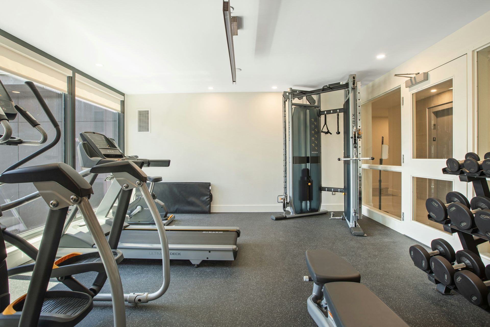 Gym with exercise equipment: treadmill, weight rack, multi-gym, weight bench, windows. Black floor. White walls.
