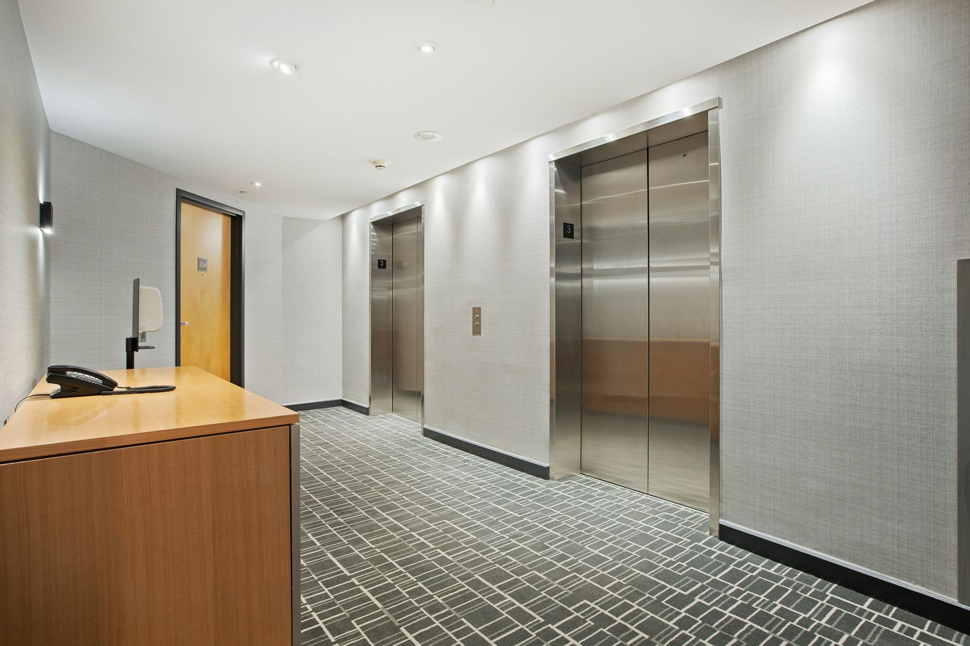 Elevator lobby with two stainless steel doors, gray walls, carpet, and wooden desk.