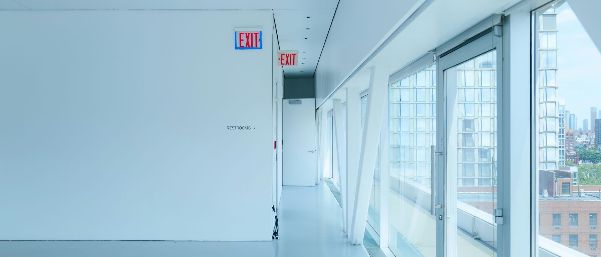Long hallway with white walls and large windows overlooking a city. Exit signs visible.