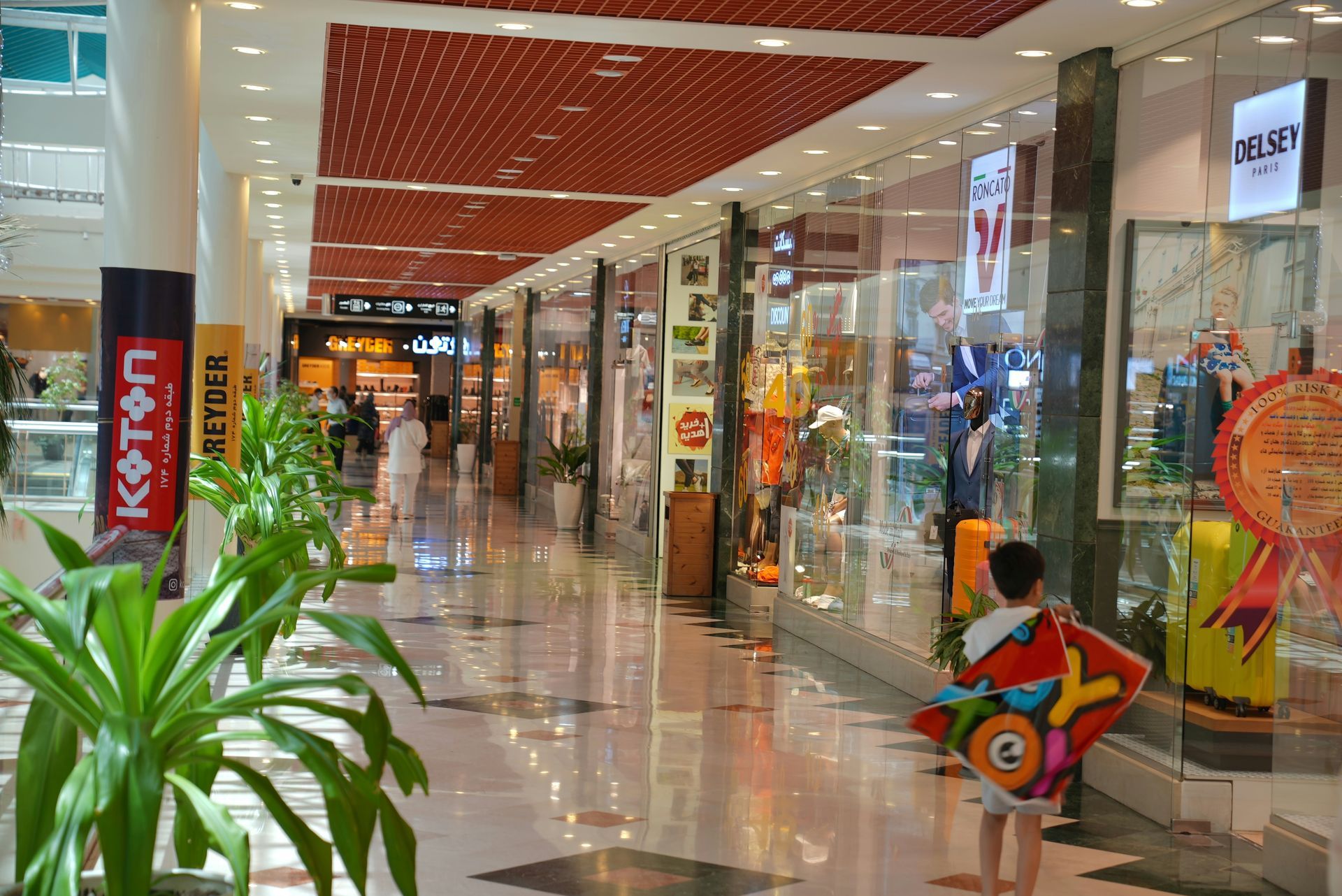 Mall interior with shops, a child holding a kite, and potted plants.