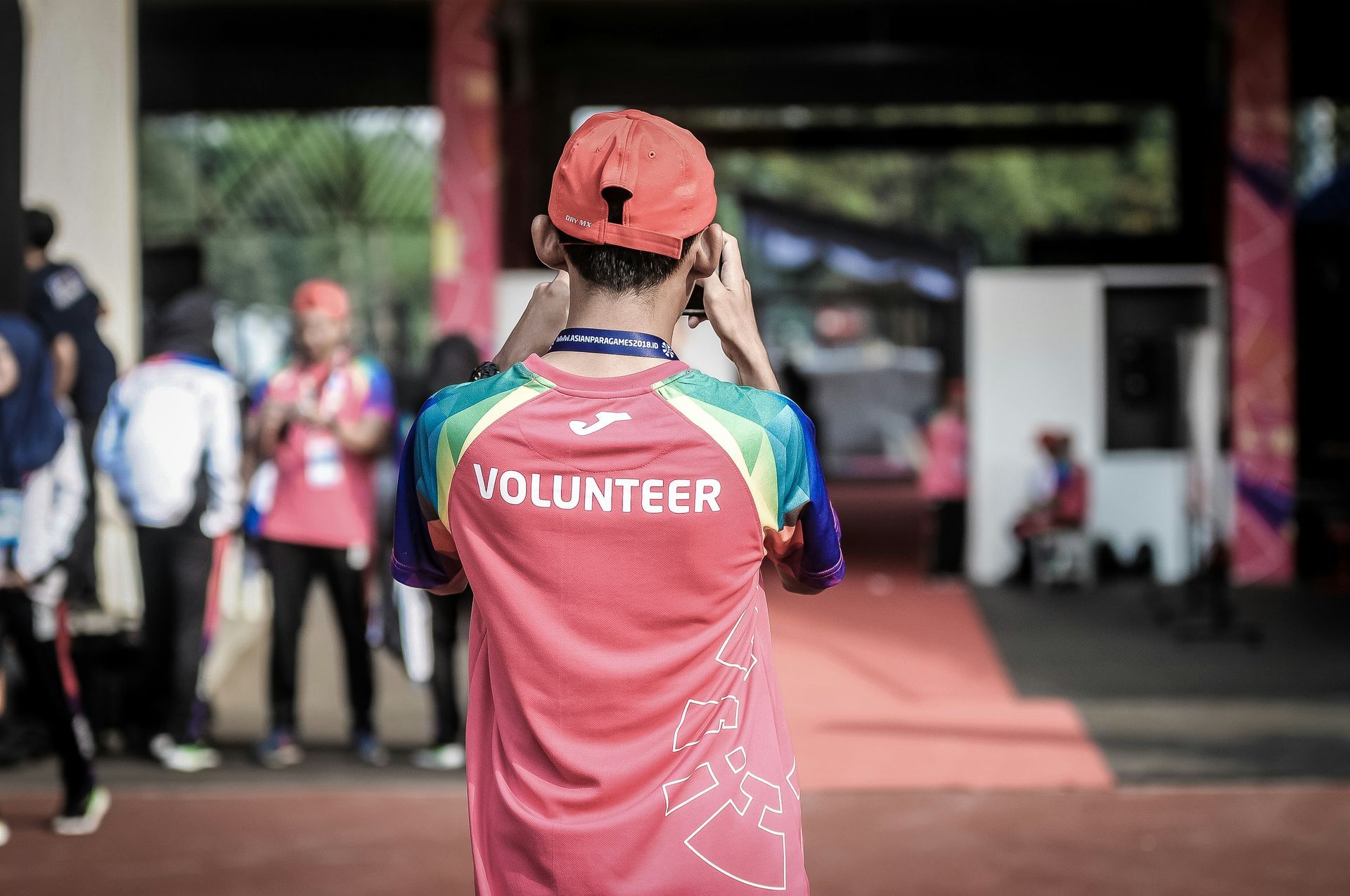 Volunteer wearing a red shirt and cap, standing outdoors at an event, observing the scene.