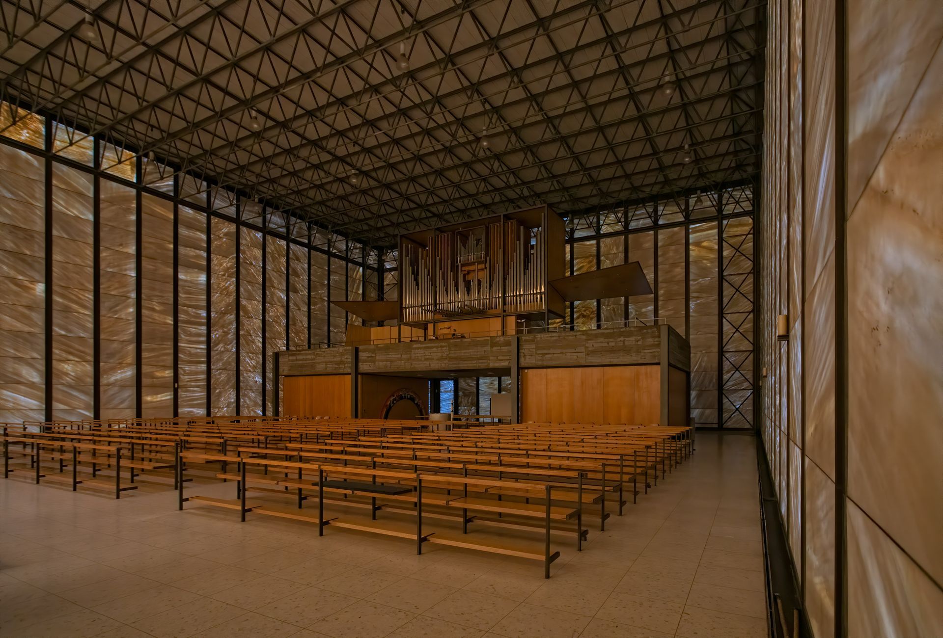 Interior of a modern church with rows of wooden pews facing an organ, walls made of glass.