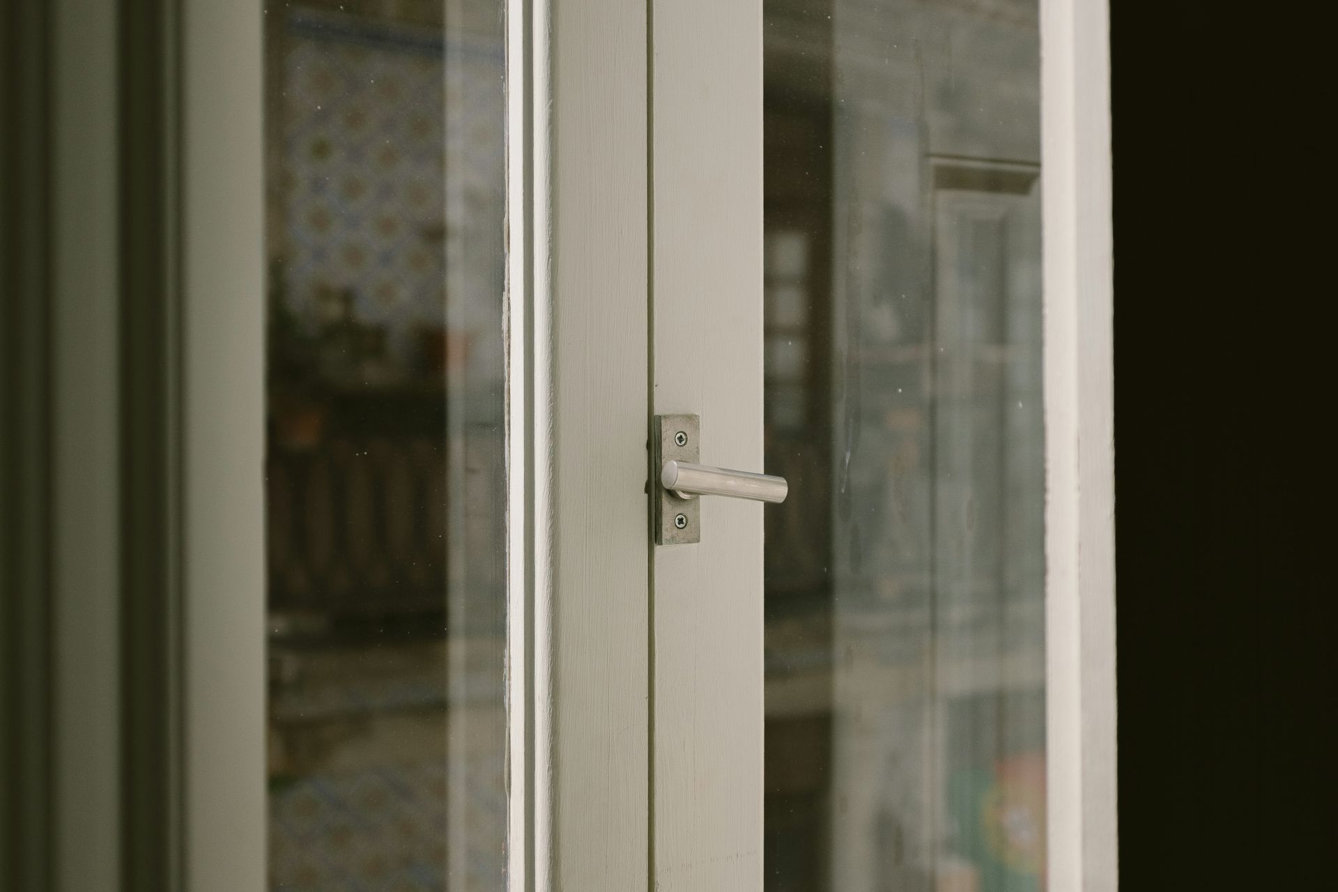 Close-up of a white window frame with a silver handle. Through the glass, a blurry interior is visible.