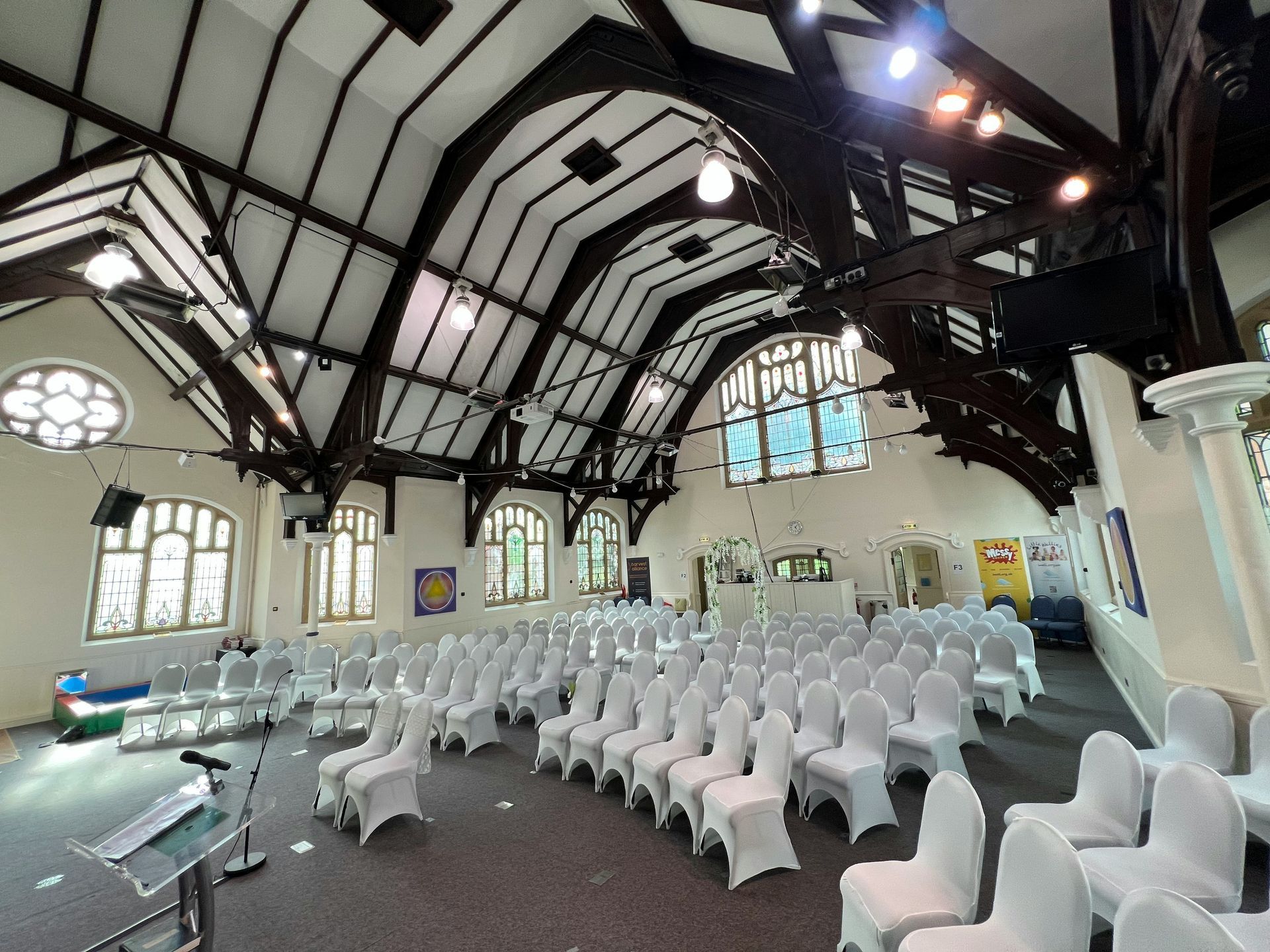 An empty auditorium with white chairs, stage, and ornate wooden beams. Daylight streams through the windows.