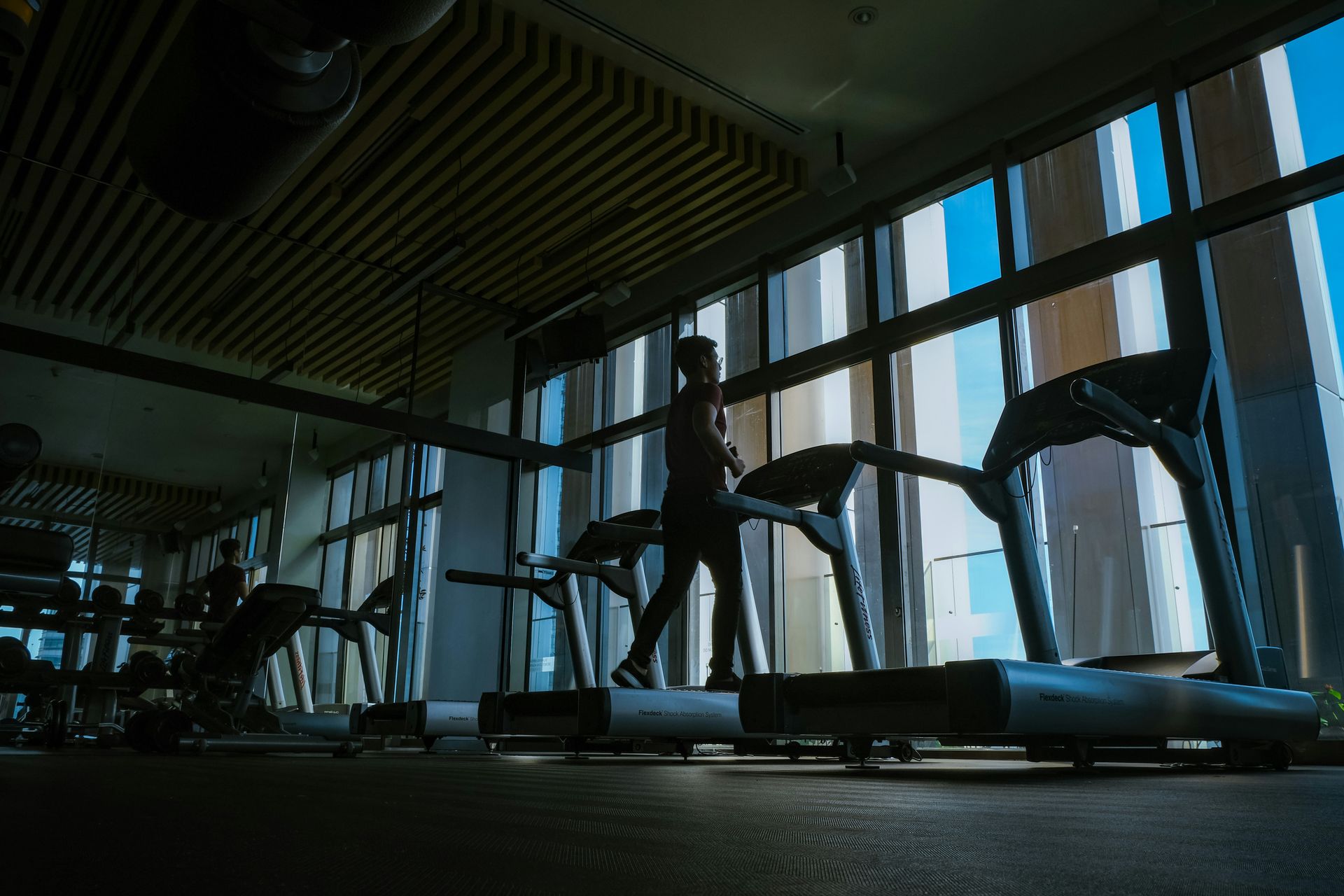 Person walking on a treadmill in a gym with large windows, bright blue sky visible.