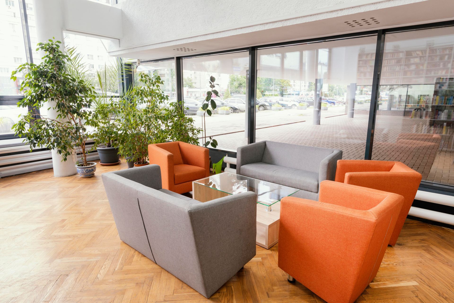 Modern office seating area with gray and orange sofas and armchairs around a glass table, with large windows and plants.
