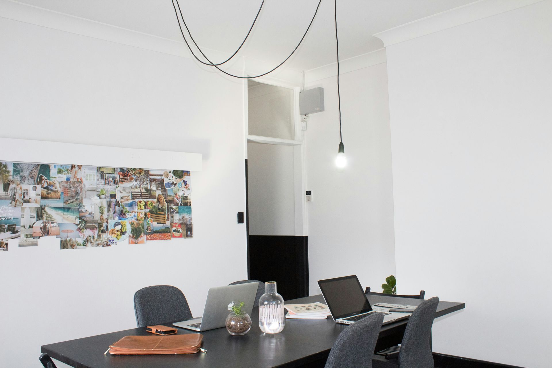 Office with a black desk, laptops, chairs, water bottle, and mood board on the wall. A hanging light illuminates the space.