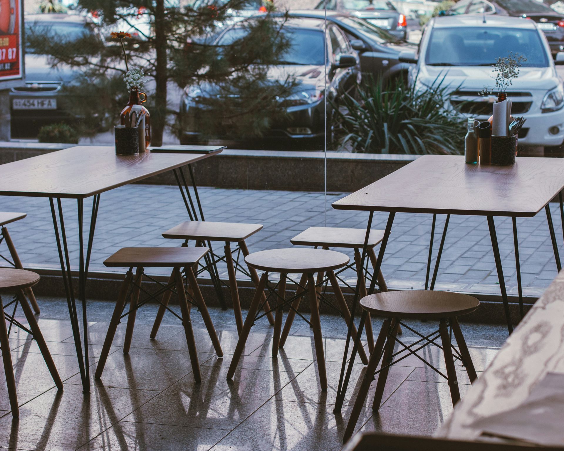 Empty outdoor cafe tables and stools; cars and greenery visible through the window.