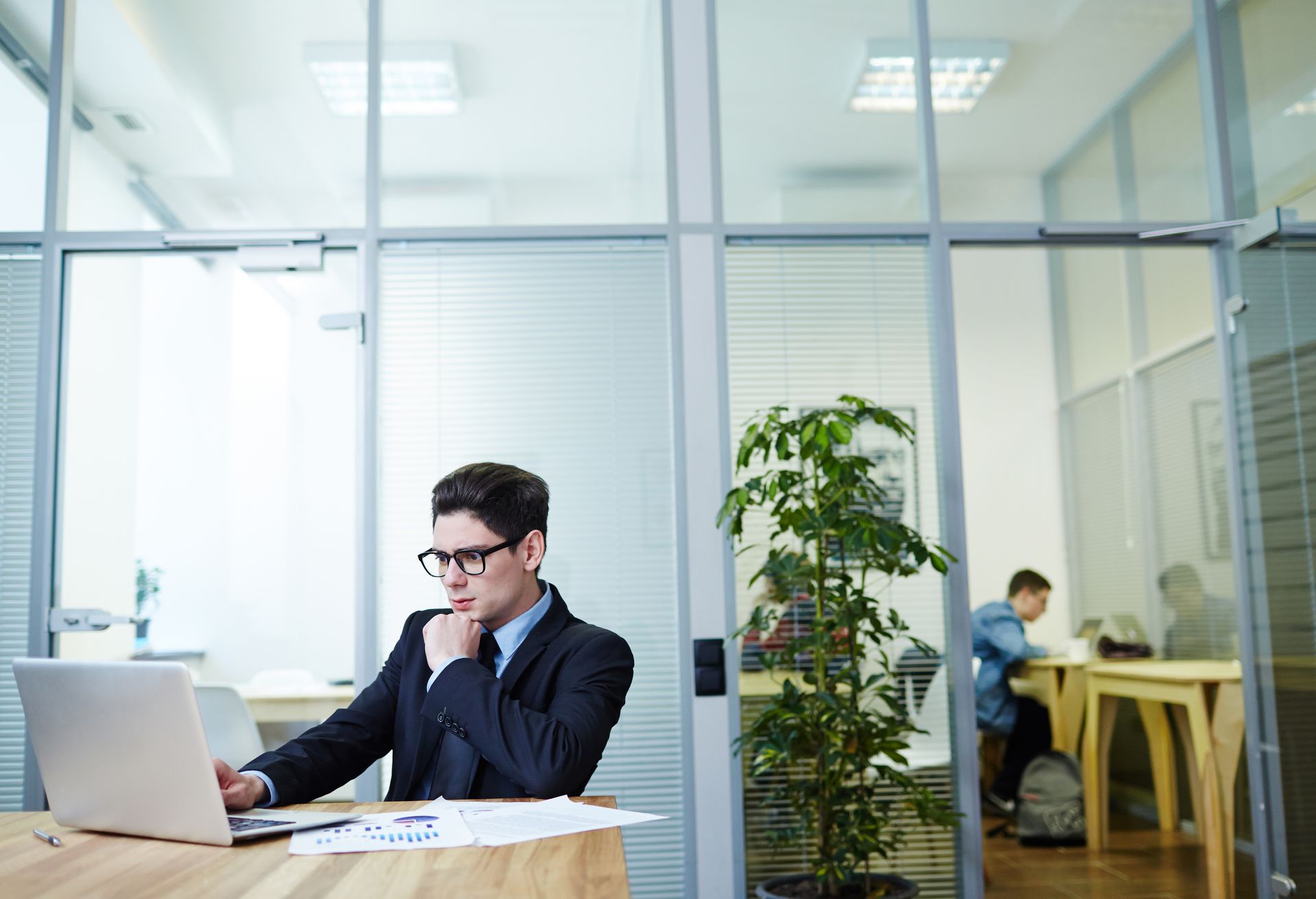 Man in suit works on laptop in office; other people visible through glass.