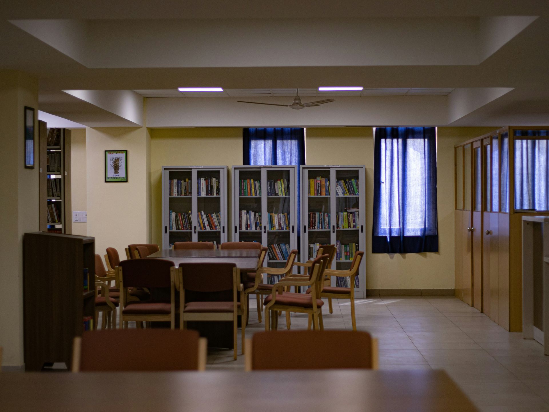 Library with bookshelves, round table, chairs, and windows with dark blue curtains.