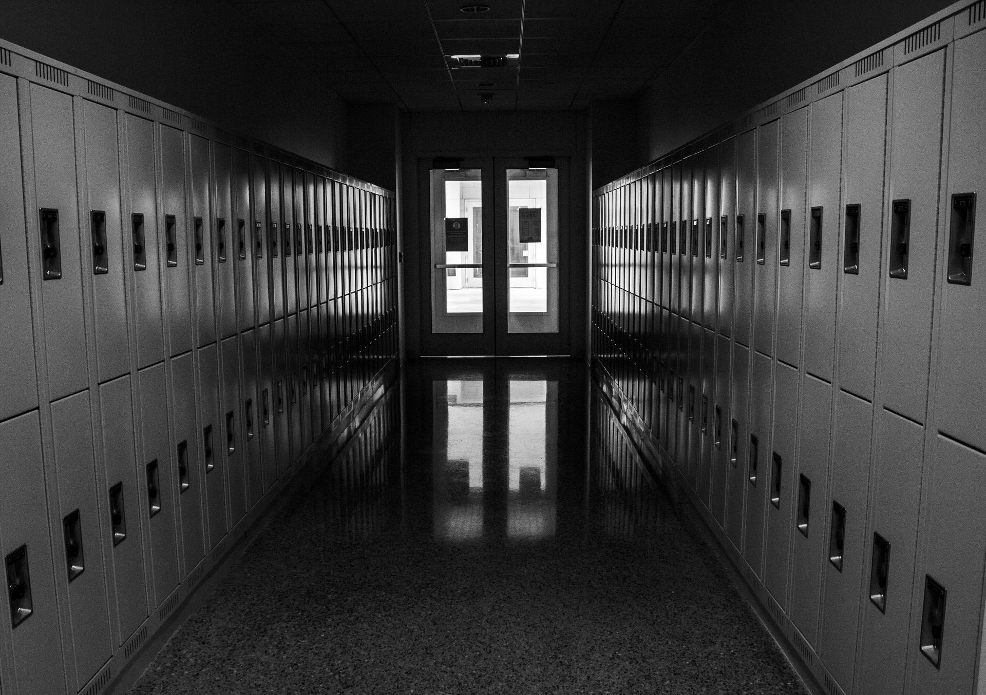 Empty school hallway with rows of lockers, doors at the end. Black and white.