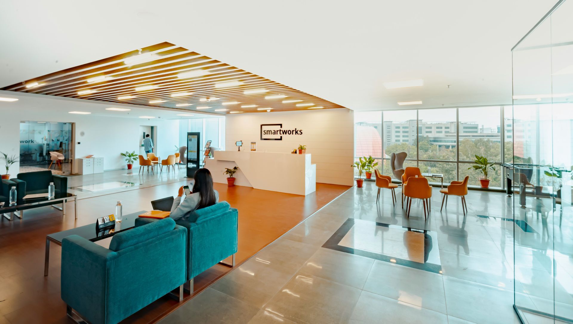 Modern office lobby with a woman on a teal couch. Bright space with orange chairs, wooden floors, and a reception desk.
