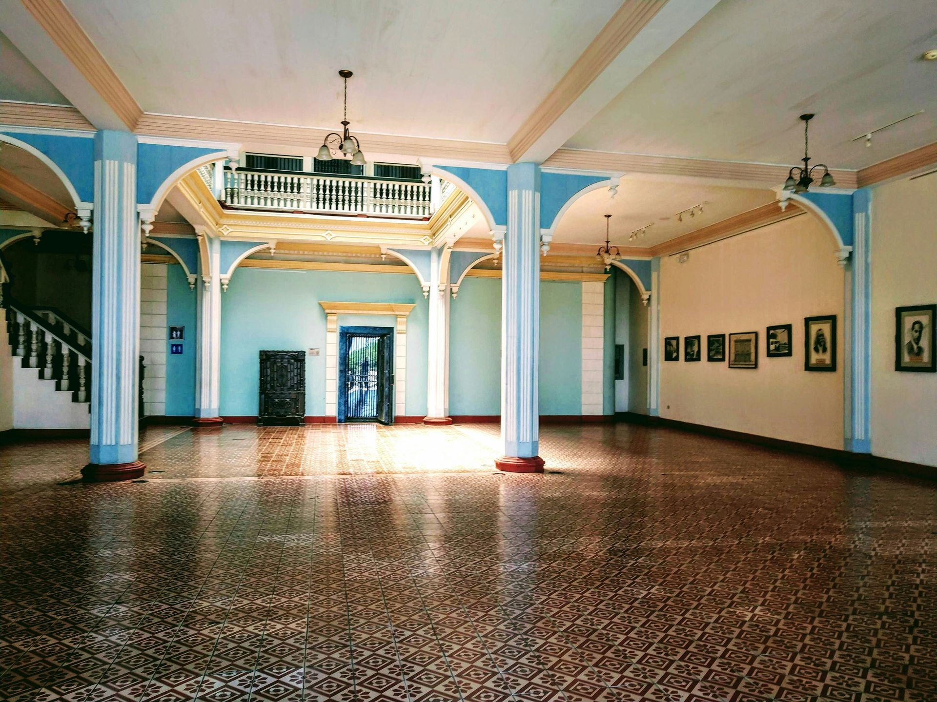 Grand hall with tiled floor, arched blue and white accents, a balcony, and portraits on the wall.