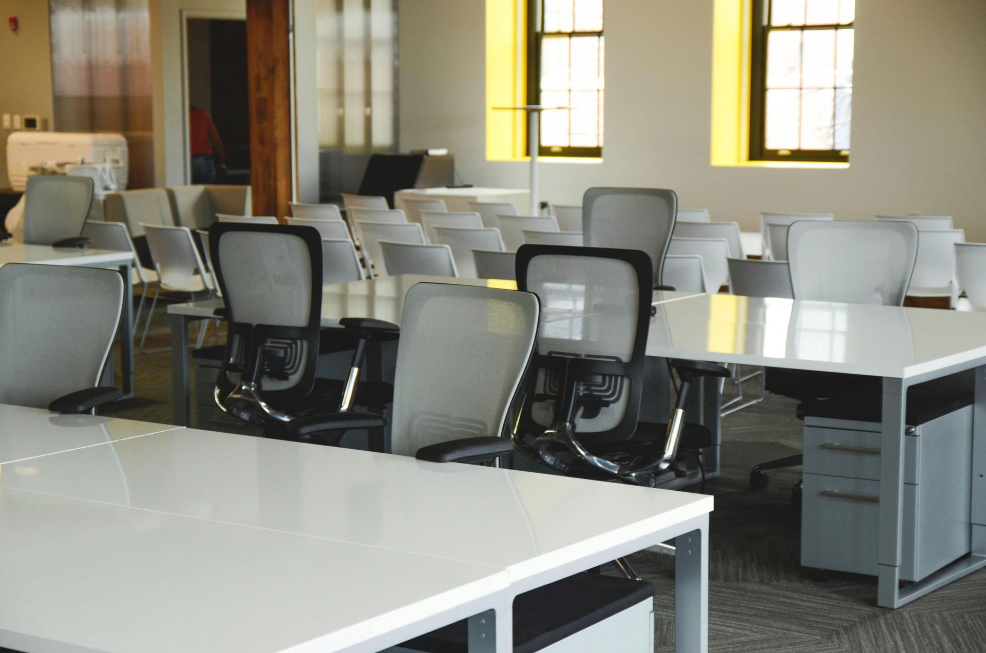 A bright, modern office space with multiple white desks, gray ergonomic chairs, and large windows.