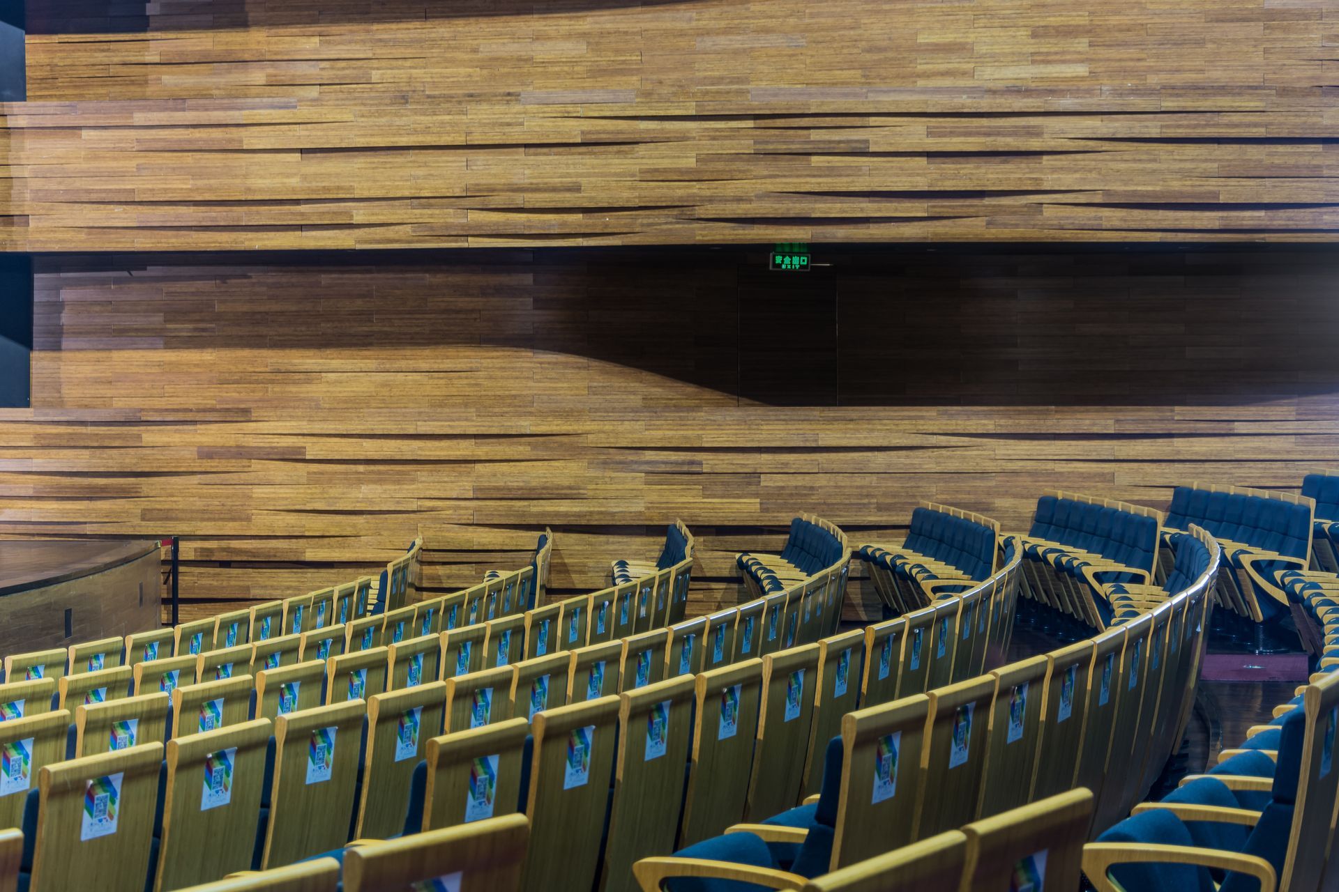 Rows of empty theater seats face a wooden wall. An illuminated exit sign hangs above.