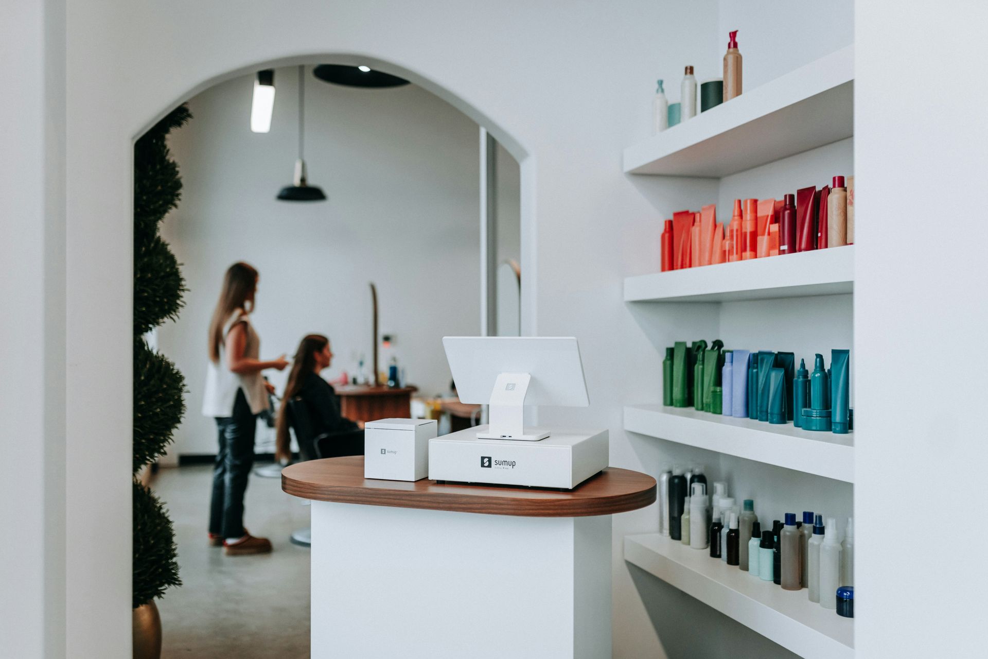 Hair salon interior with stylist working on client, display shelves with product, and a checkout counter.