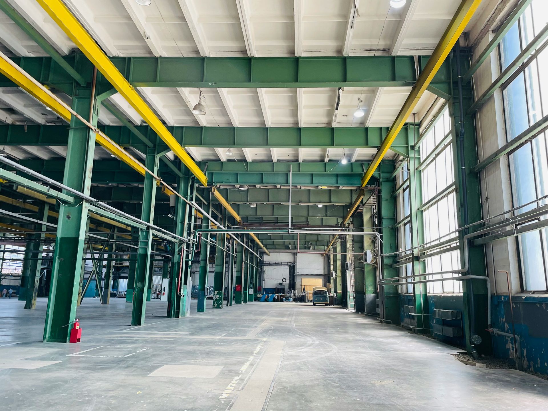 Empty industrial warehouse with green support beams, yellow overhead pipes, and large windows.
