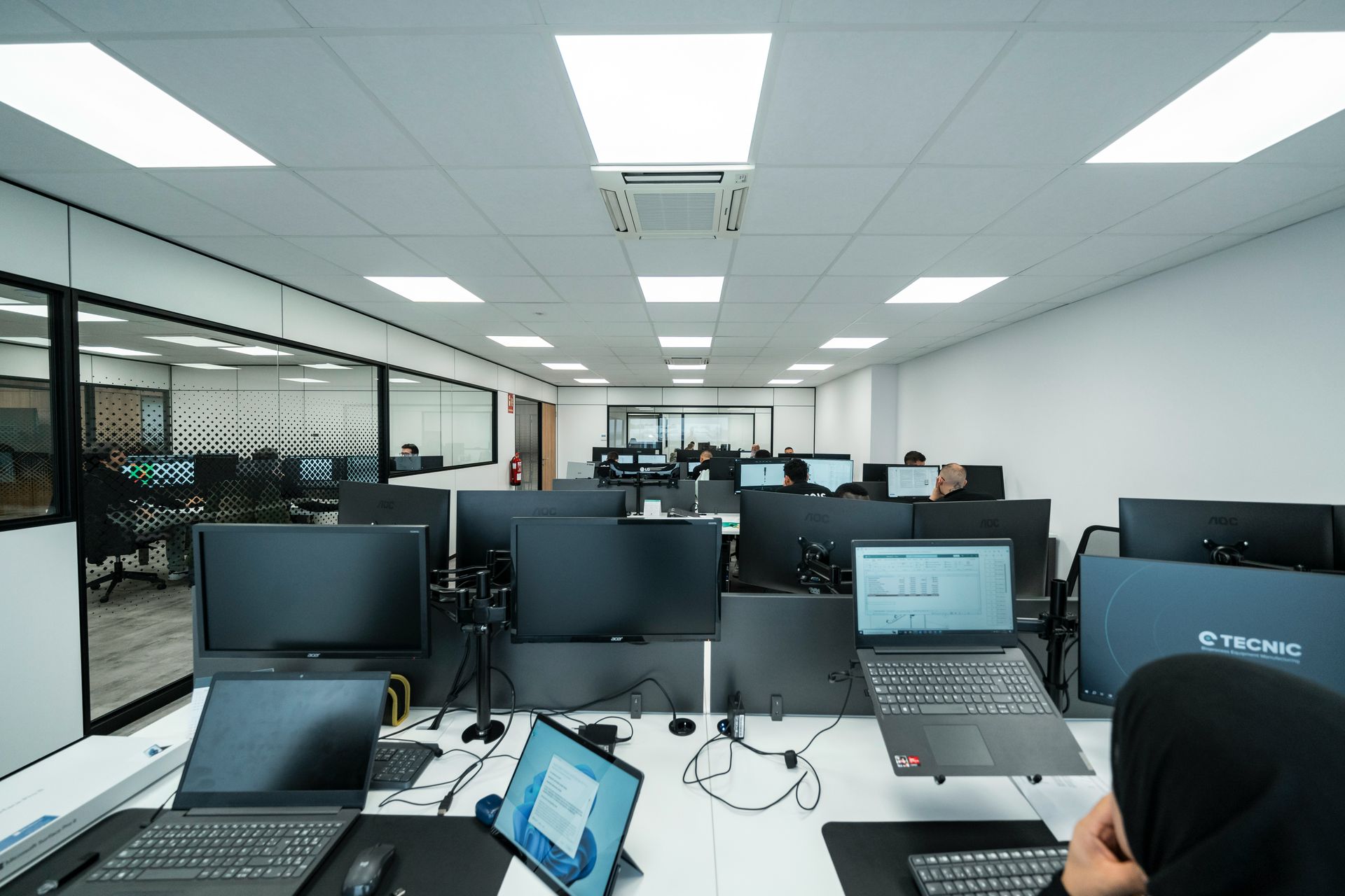 An office space with computers on desks. People are working. Fluorescent lights shine from the ceiling.