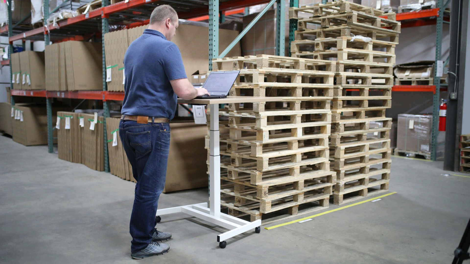Man using a laptop at a standing desk in a warehouse; cardboard boxes and pallets visible.