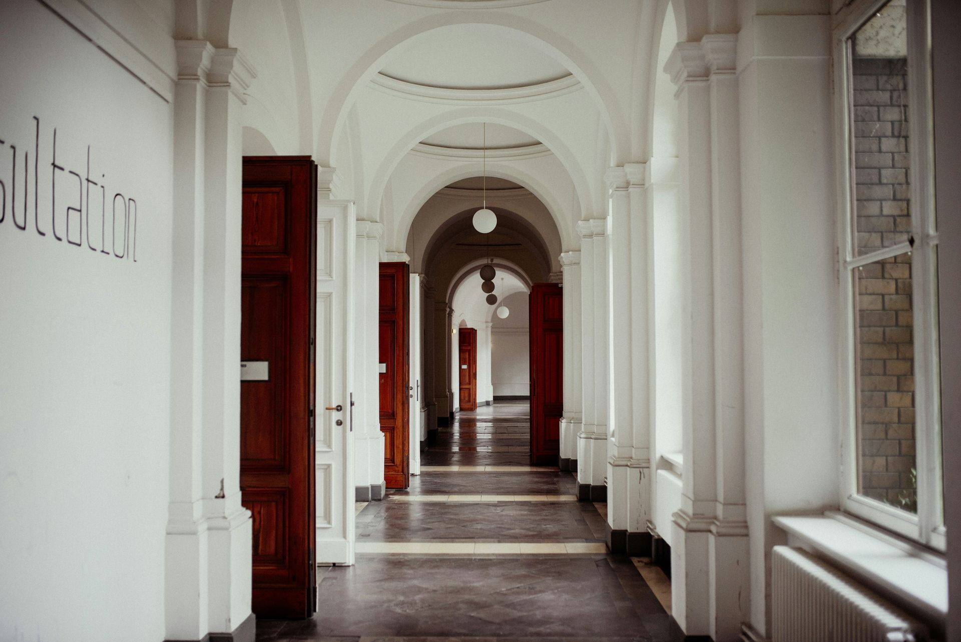 Long hallway with white arched ceiling and columns, brown doors, and a window.