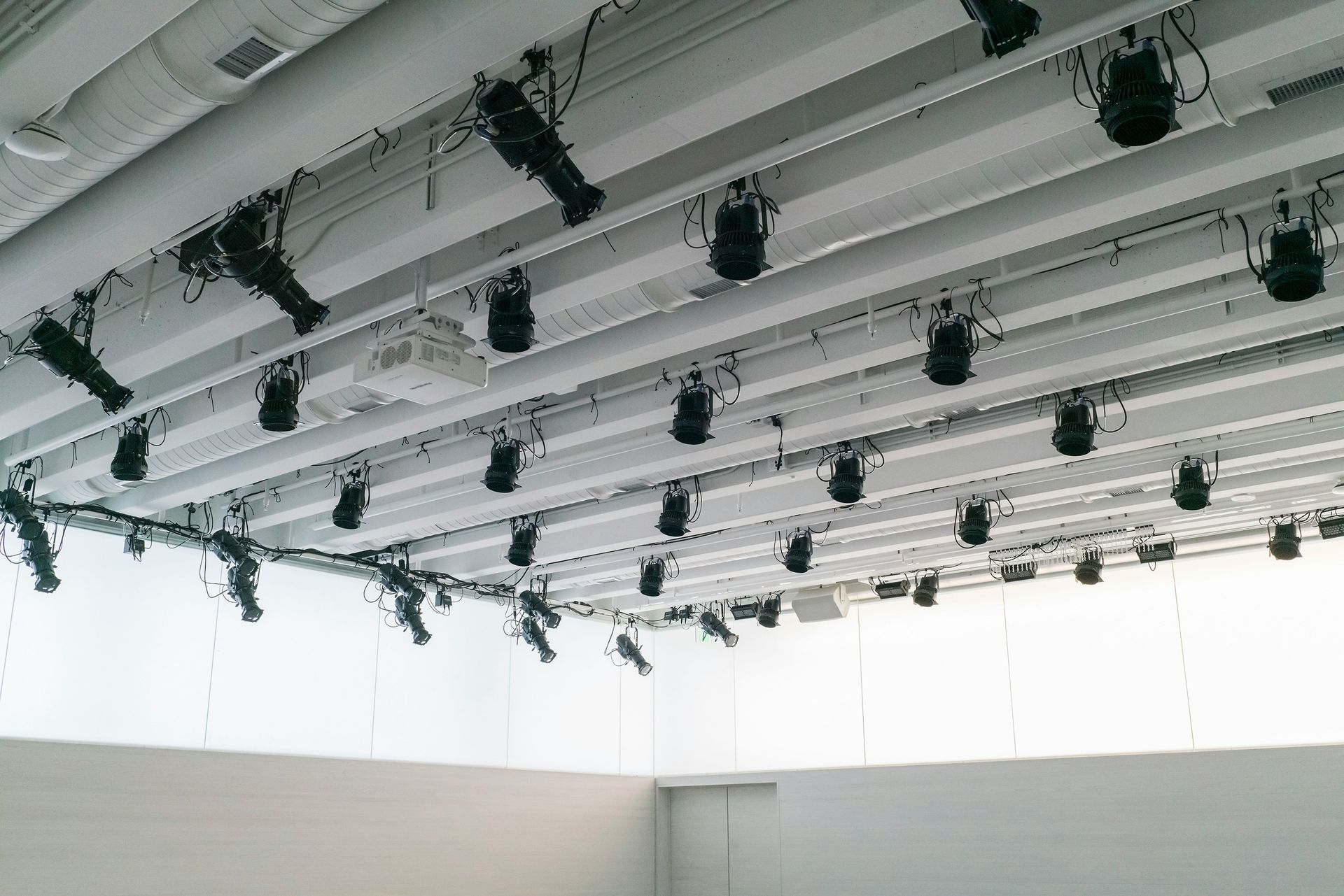 Rows of stage lights hang from a white ceiling, angled towards a blank, white room.