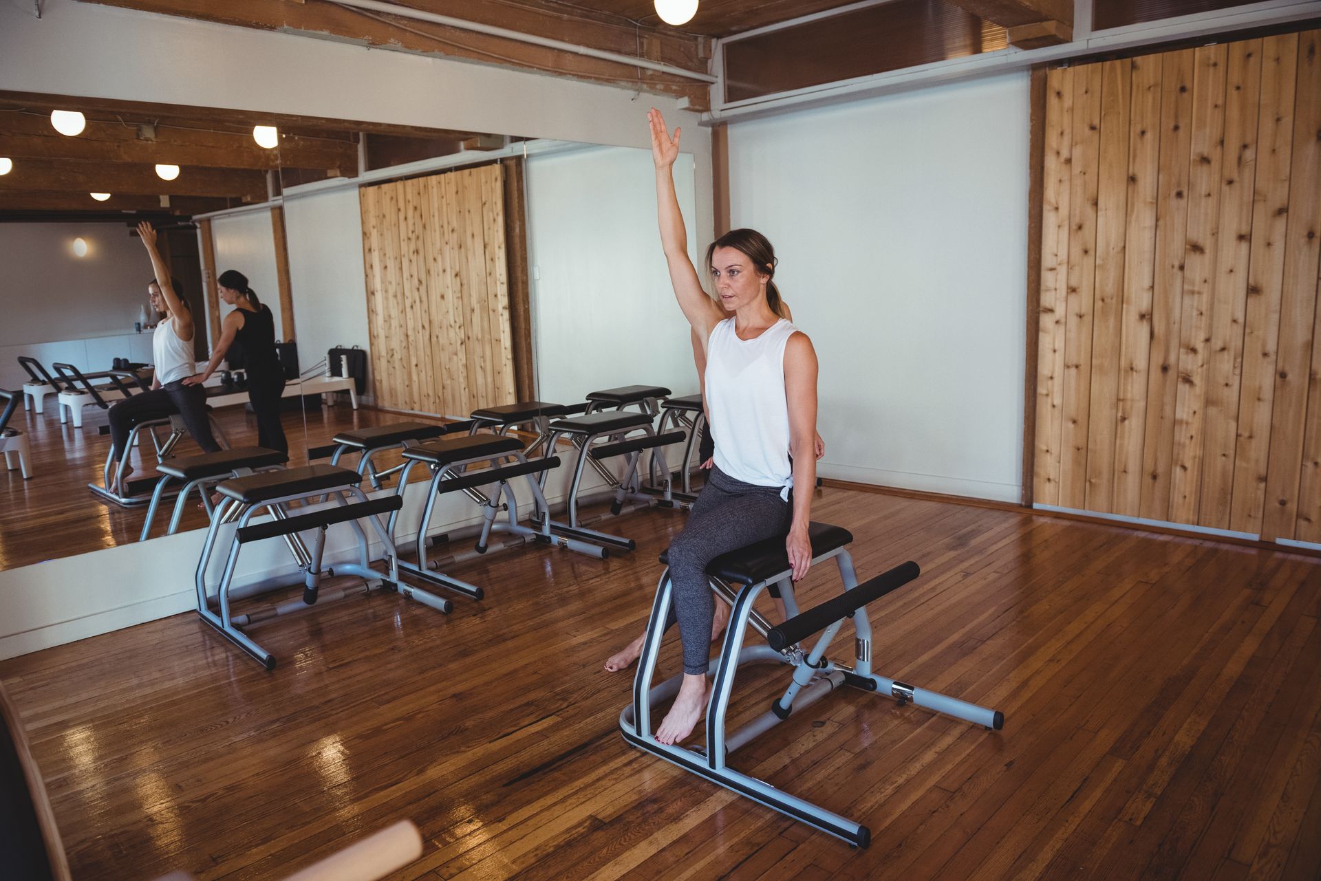 Woman in white top exercises on Pilates chair, arm raised, in a studio with wooden floor and mirrors.