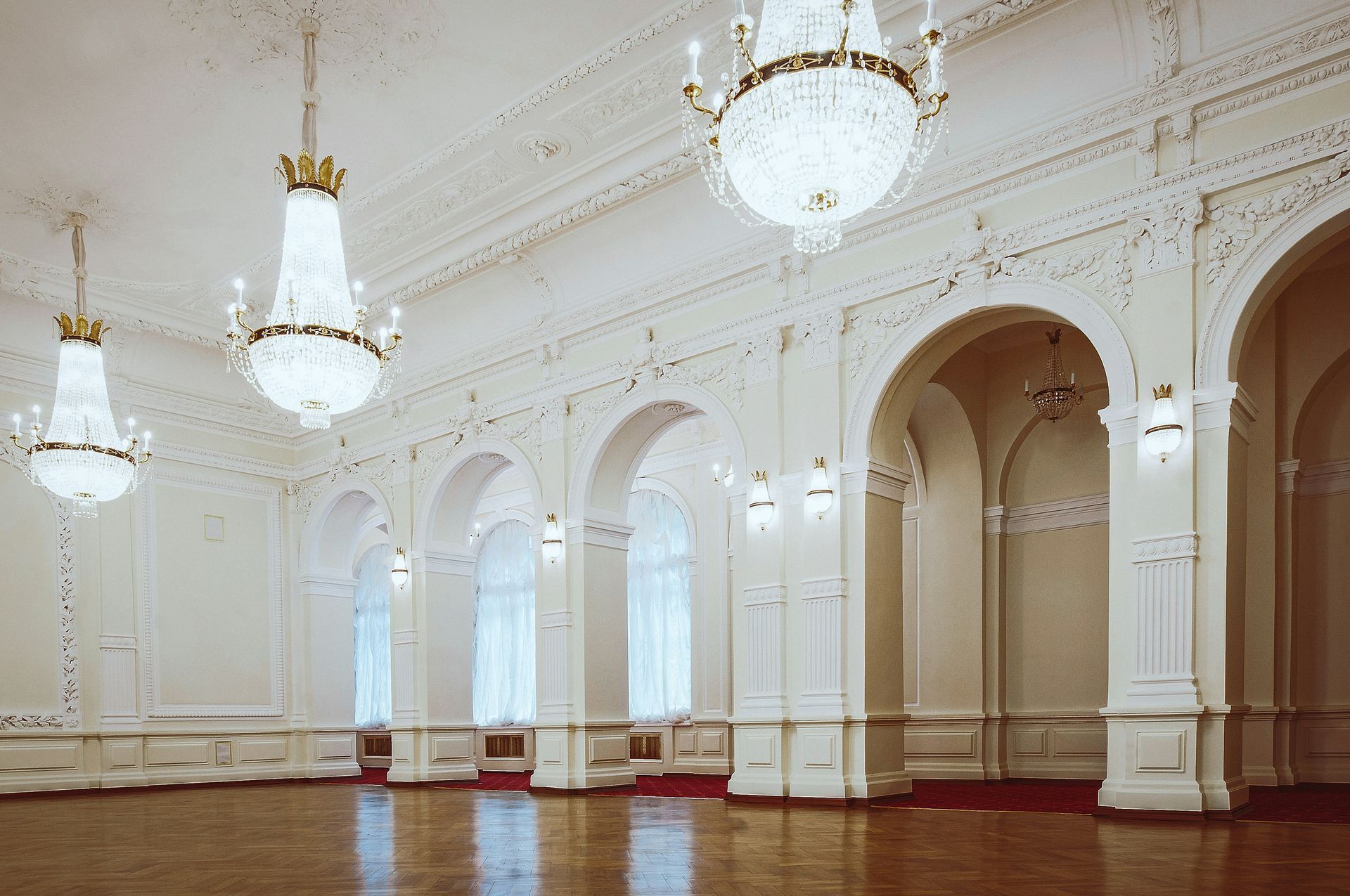 Ornate white ballroom with arched windows, crystal chandeliers, and wooden floor.