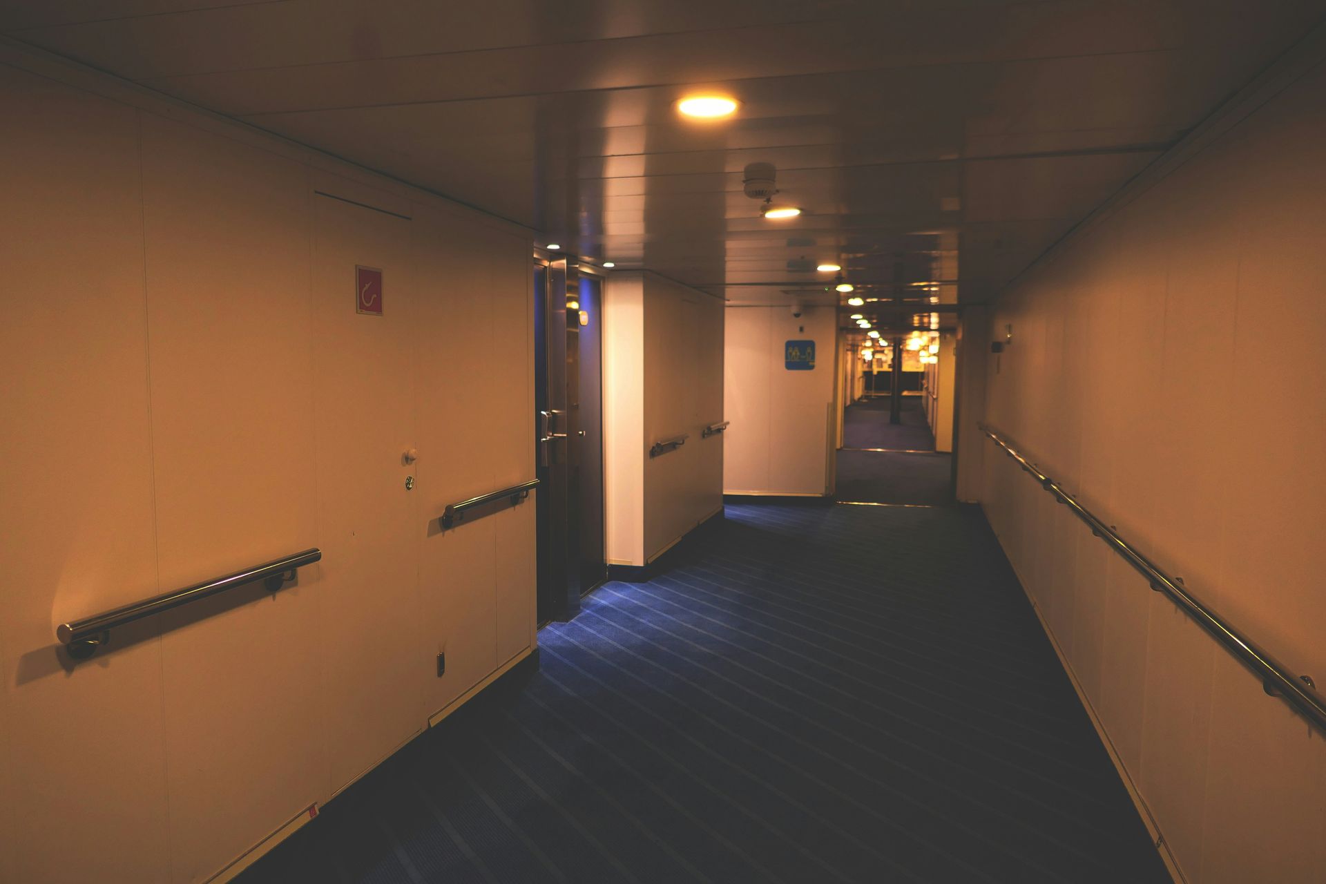 Long, dimly lit hallway with blue carpet, white walls, and handrails.