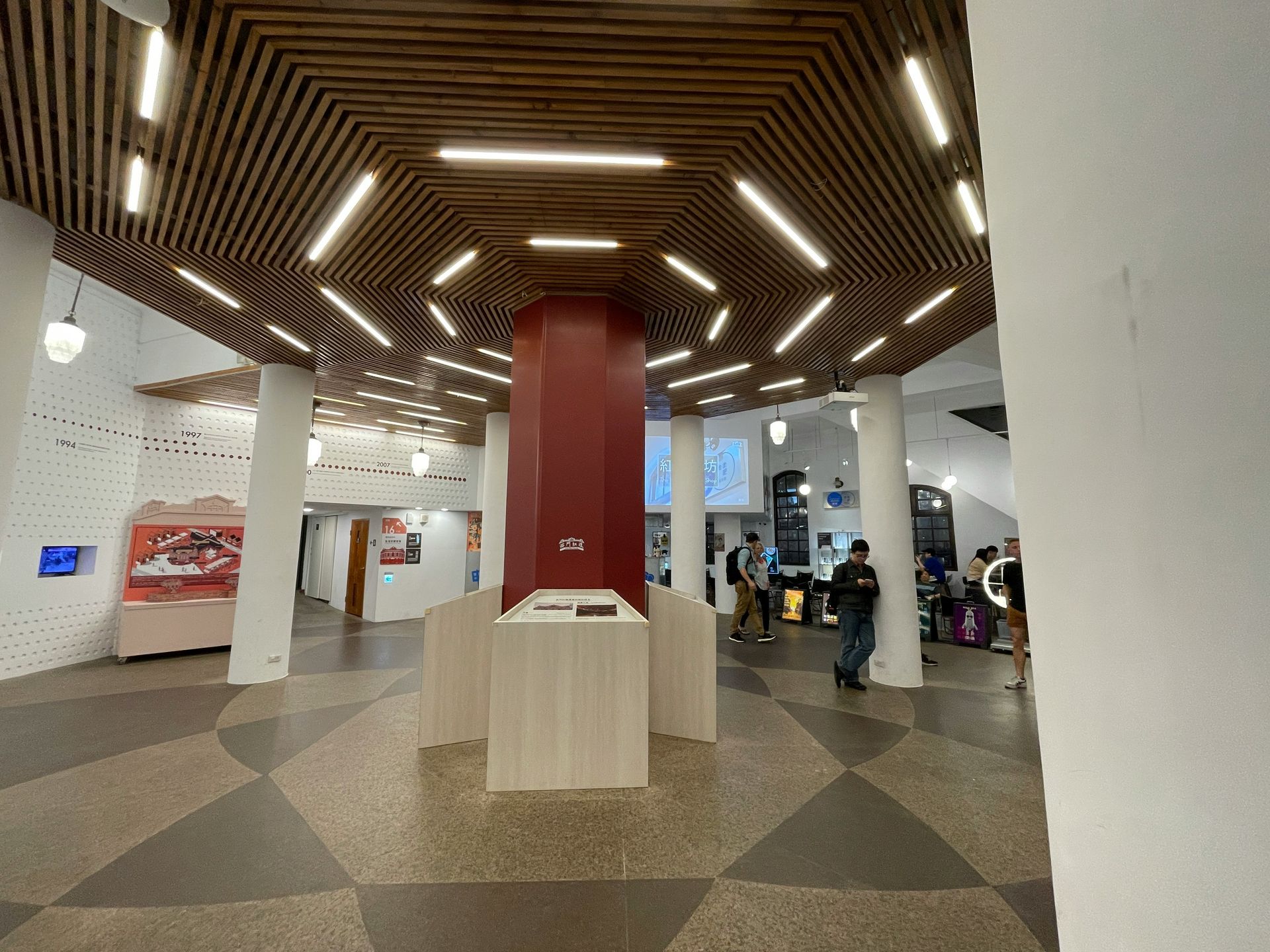Interior with a hexagonal ceiling design with lights. A red column in the center, people walking.