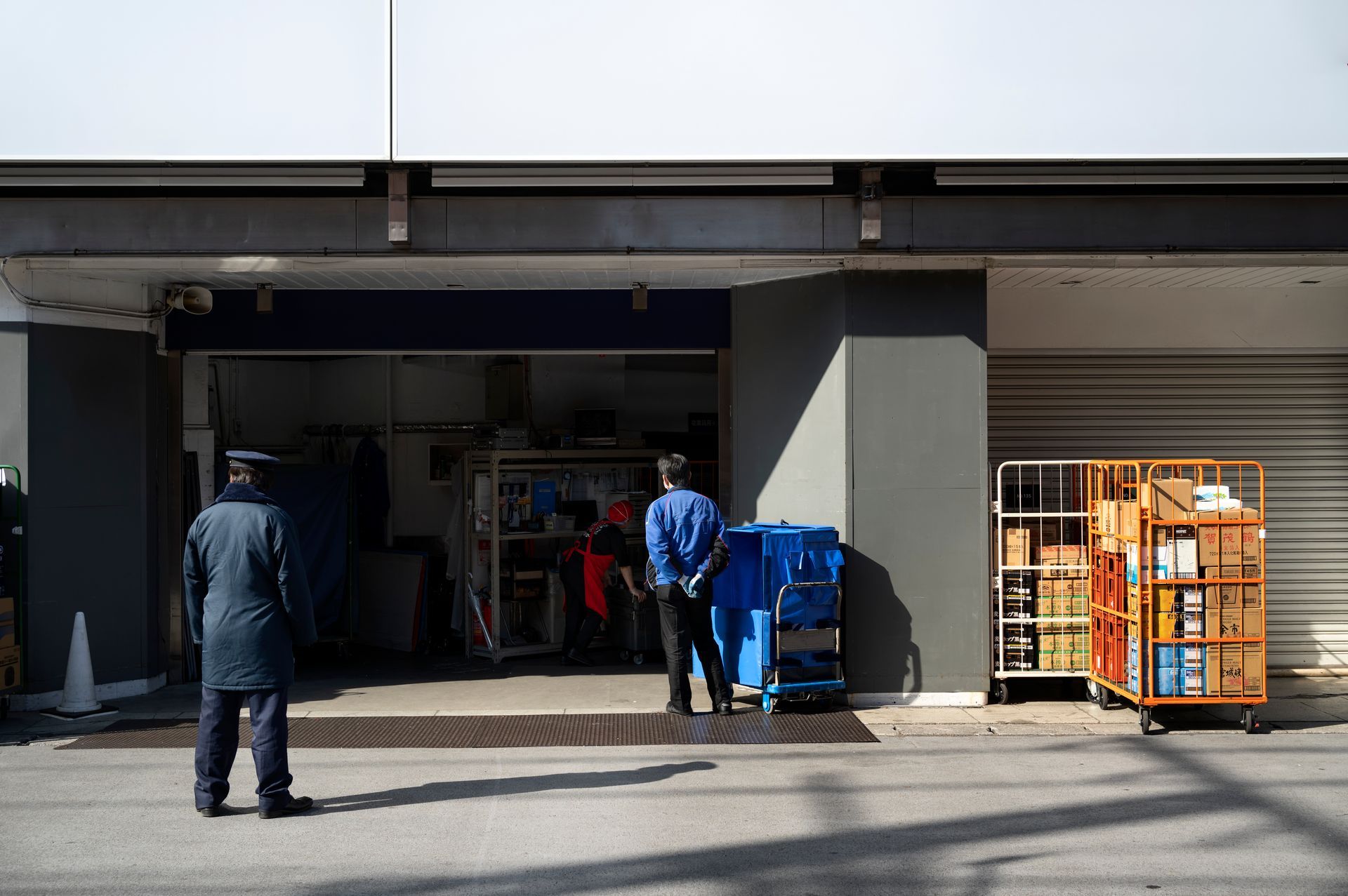 People loading a blue cart and observing a metal cage cart, outside a building entrance.