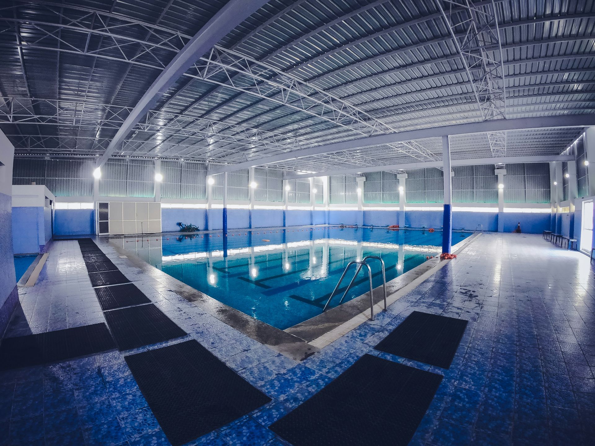 Indoor swimming pool with blue water and lane markers. Empty, with blue walls and a metal ceiling.