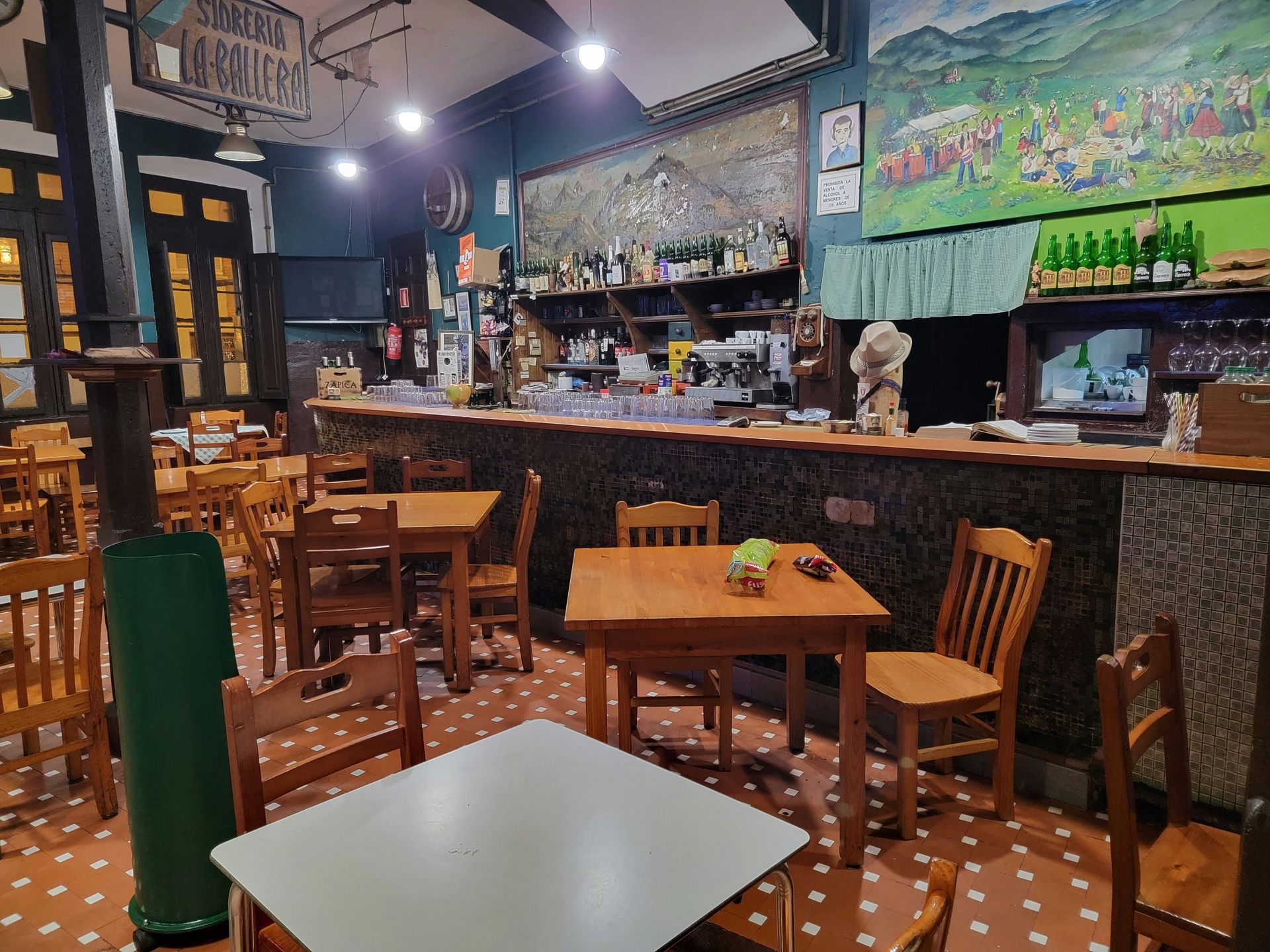 Interior of a rustic bar with wooden tables, chairs, and a bar counter. Bottles, art, and lights are visible.