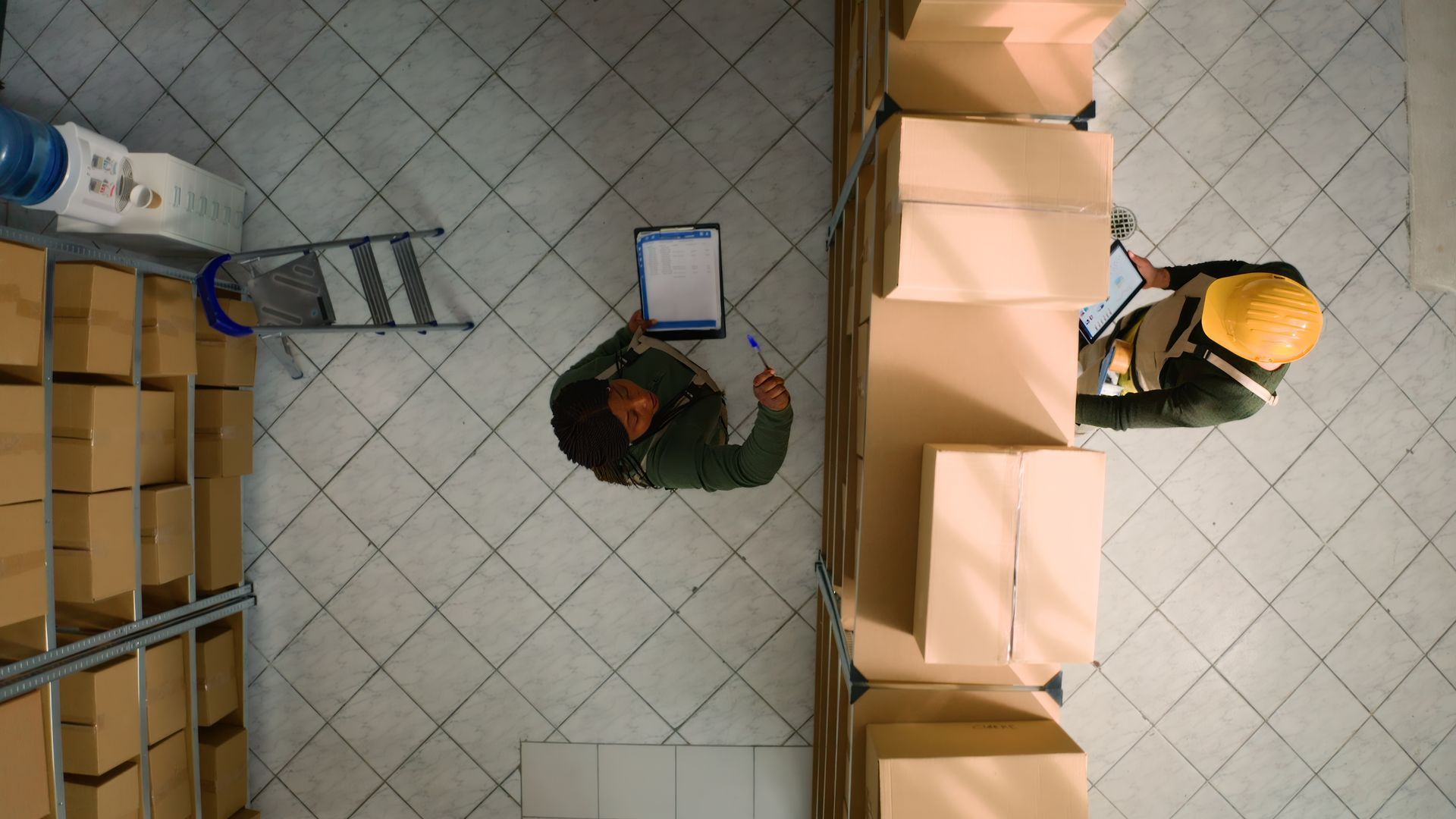 Overhead view of two workers in a warehouse, one with a clipboard, the other wearing a hard hat, surrounded by boxes.