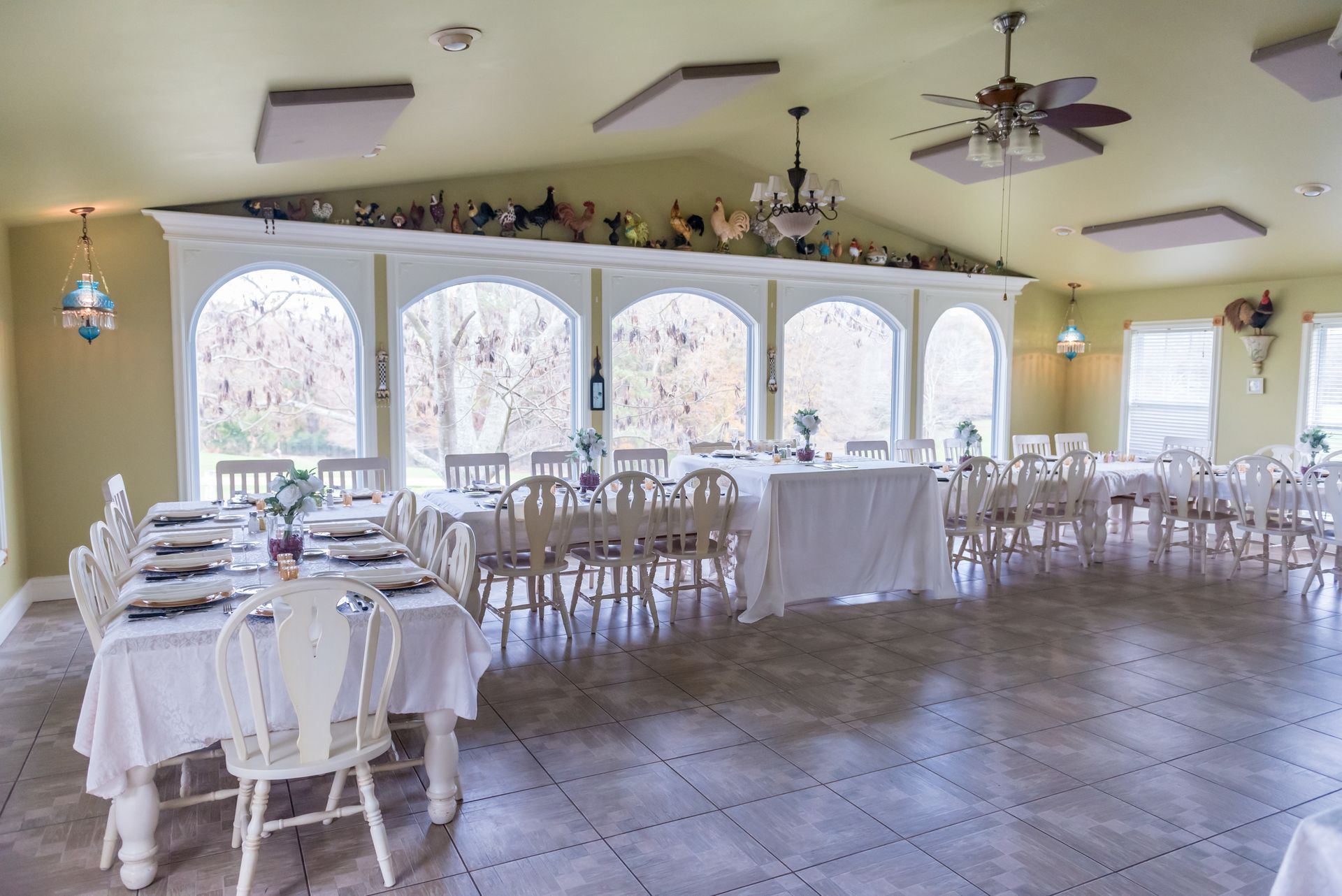 Empty dining hall with white tables and chairs, large arched windows, and decorative items.