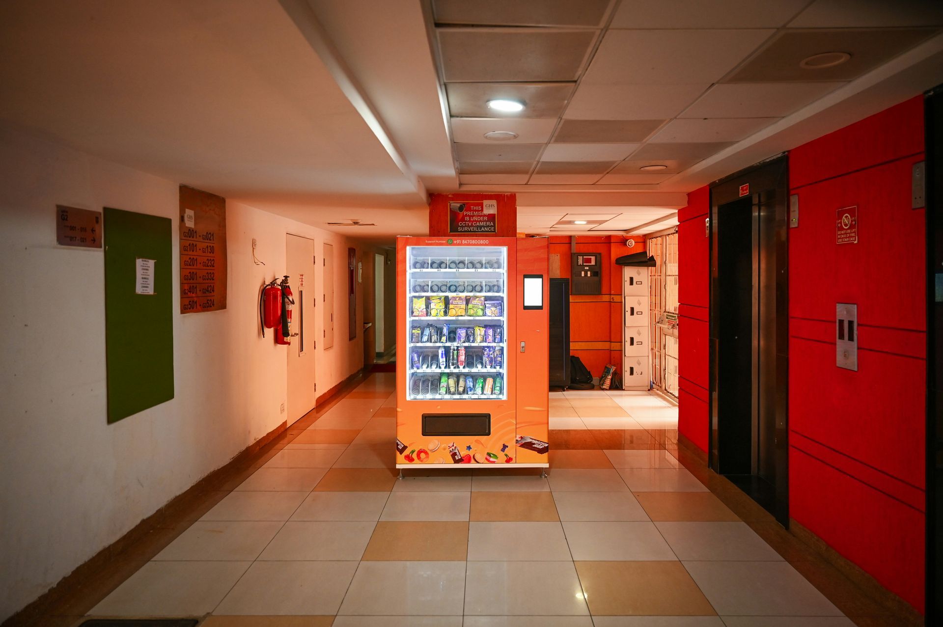 Hallway with a vending machine, elevators, and doors. Red and orange walls, tan floor tiles.