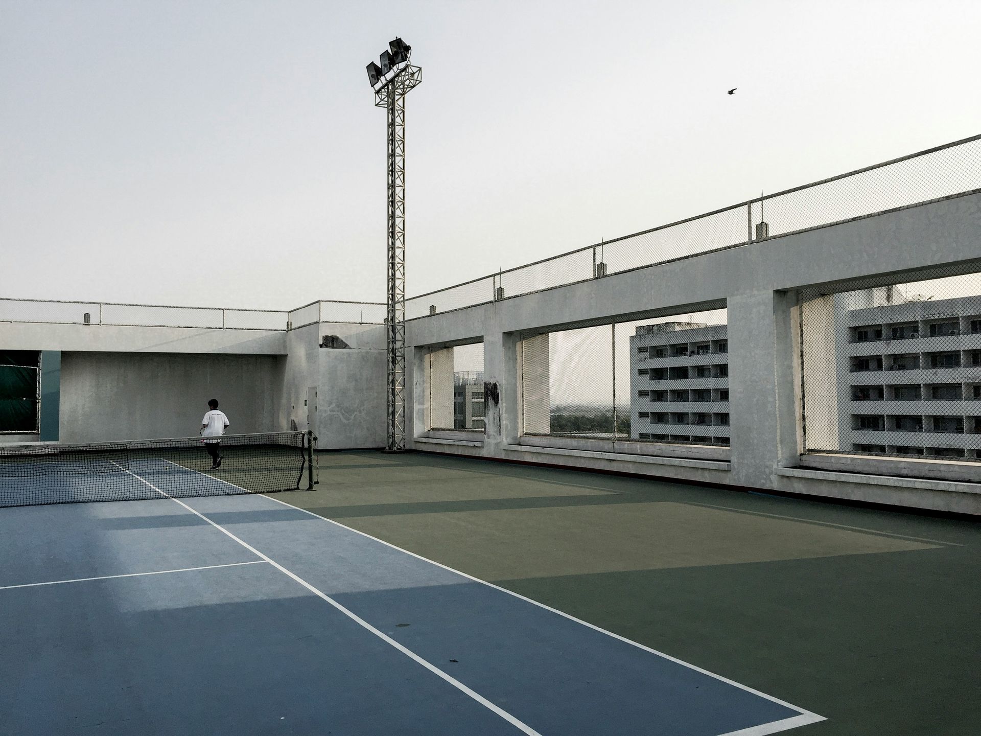 Tennis court on a rooftop with a person playing. Blue and green court, net, high fence, light pole, building background.
